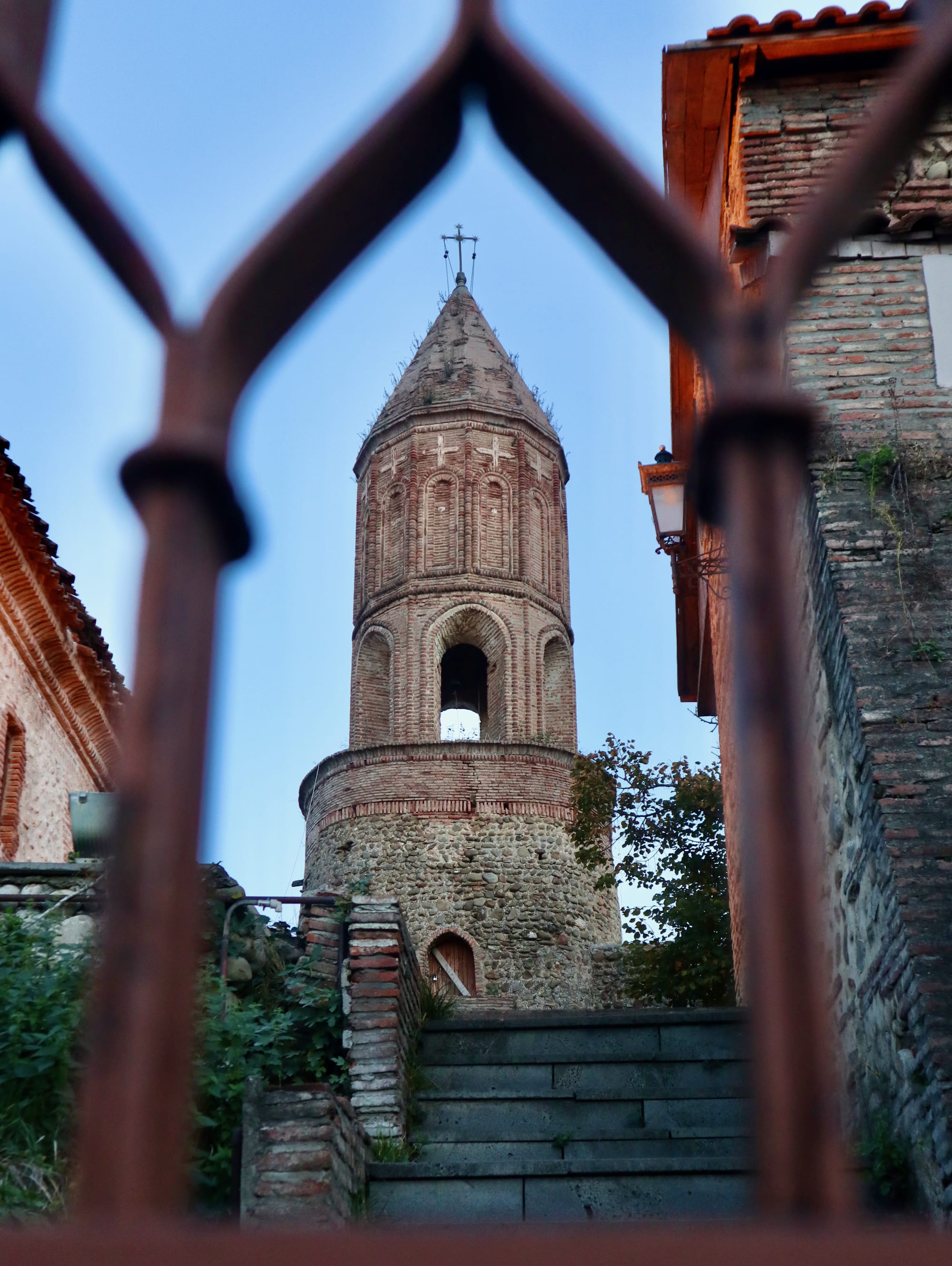 A glimpse of historic beauty unfolds as you explore the charming town of Sighnaghi, Georgia, known as the famed City of Love. As you wander through its cobblestone streets, you'll encounter a striking scene: a stone tower with an arched top viewed through a decorative metal gate, flanked by brick buildings and accompanied by a flight of stairs. This picturesque setting is just one example of the many things to do in Sighnaghi.