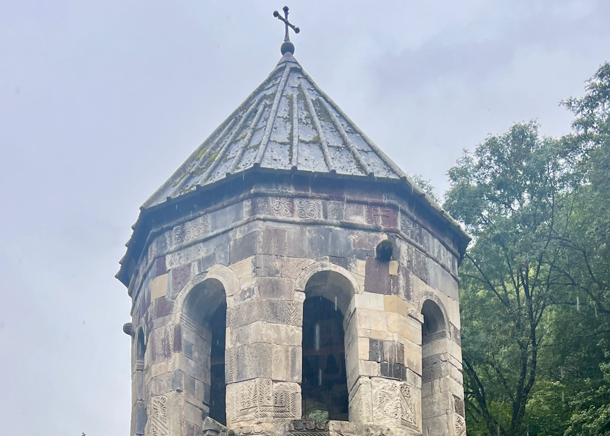 Amidst the natural beauty of Borjomi, a stone tower with arched windows and a metal cross atop stands quietly, surrounded by trees under a cloudy sky, echoing the region's rich culture. This serene setting makes one ponder: is Borjomi worth visiting for those seeking both scenic tranquility and a touch of cultural heritage?