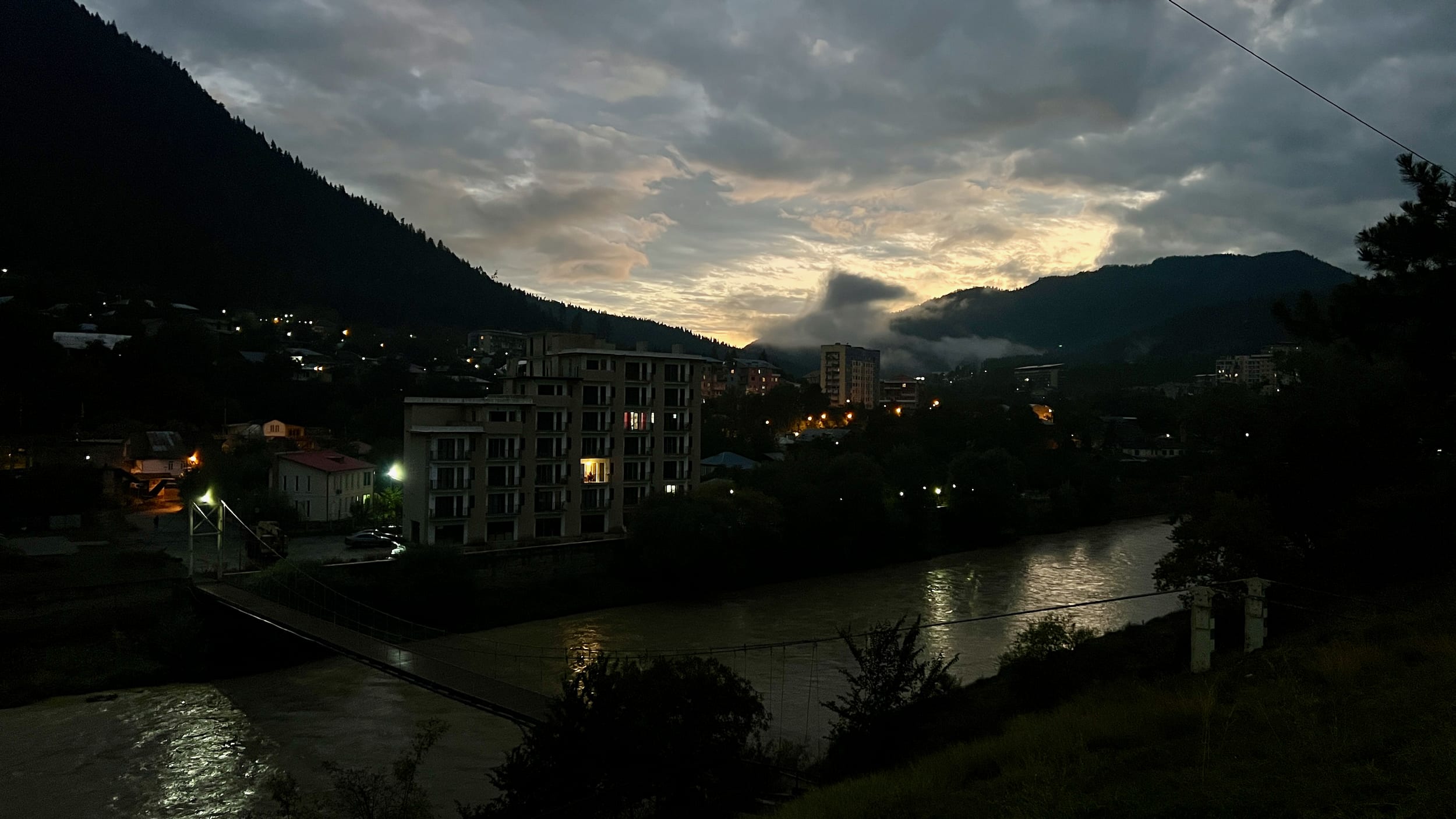 Borjomi at dusk unveils its beauty with silhouetted hills against a cloudy sky, illuminated buildings, and a river reflecting the dim light. With such stunning scenery on offer, one might wonder, is Borjomi worth visiting? The answer lies in its enchanting landscapes and inviting ambiance—a must-see spot for any traveler.