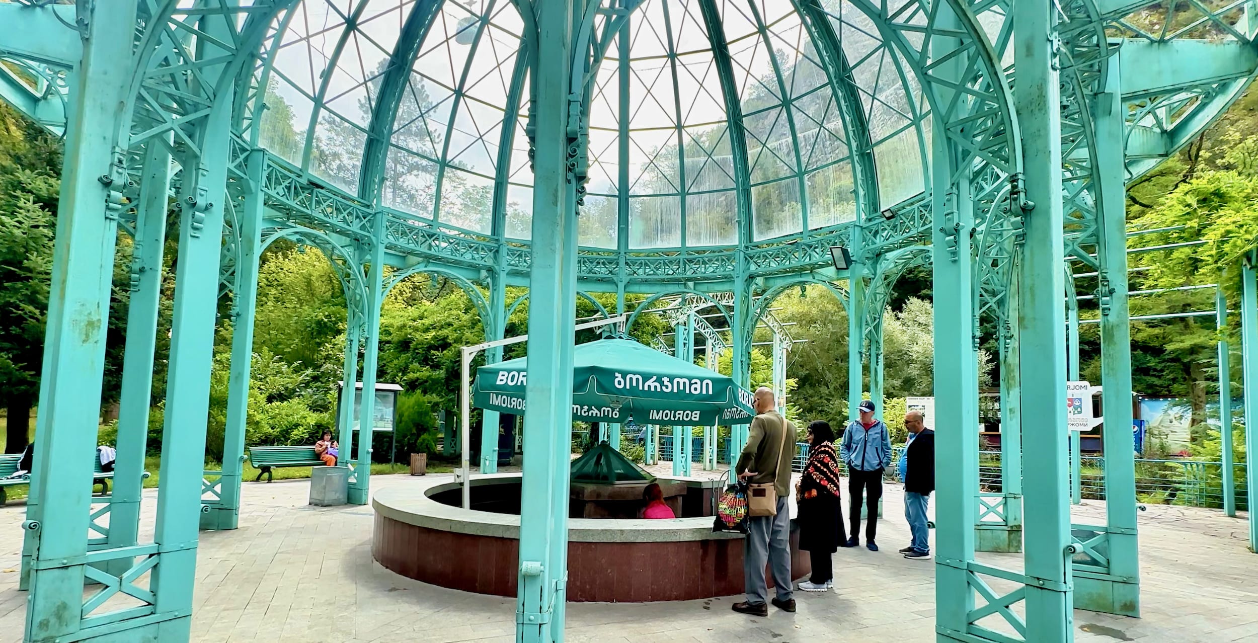Beneath a turquoise metal and glass pavilion, people gather around a mineral water fountain that echoes Borjomi's vibrant culture with its geometric design. As trees sway gracefully in the background, you might wonder, is Borjomi worth visiting? This captivating blend of natural beauty and cultural charm provides more than enough reason to explore this enchanting destination.