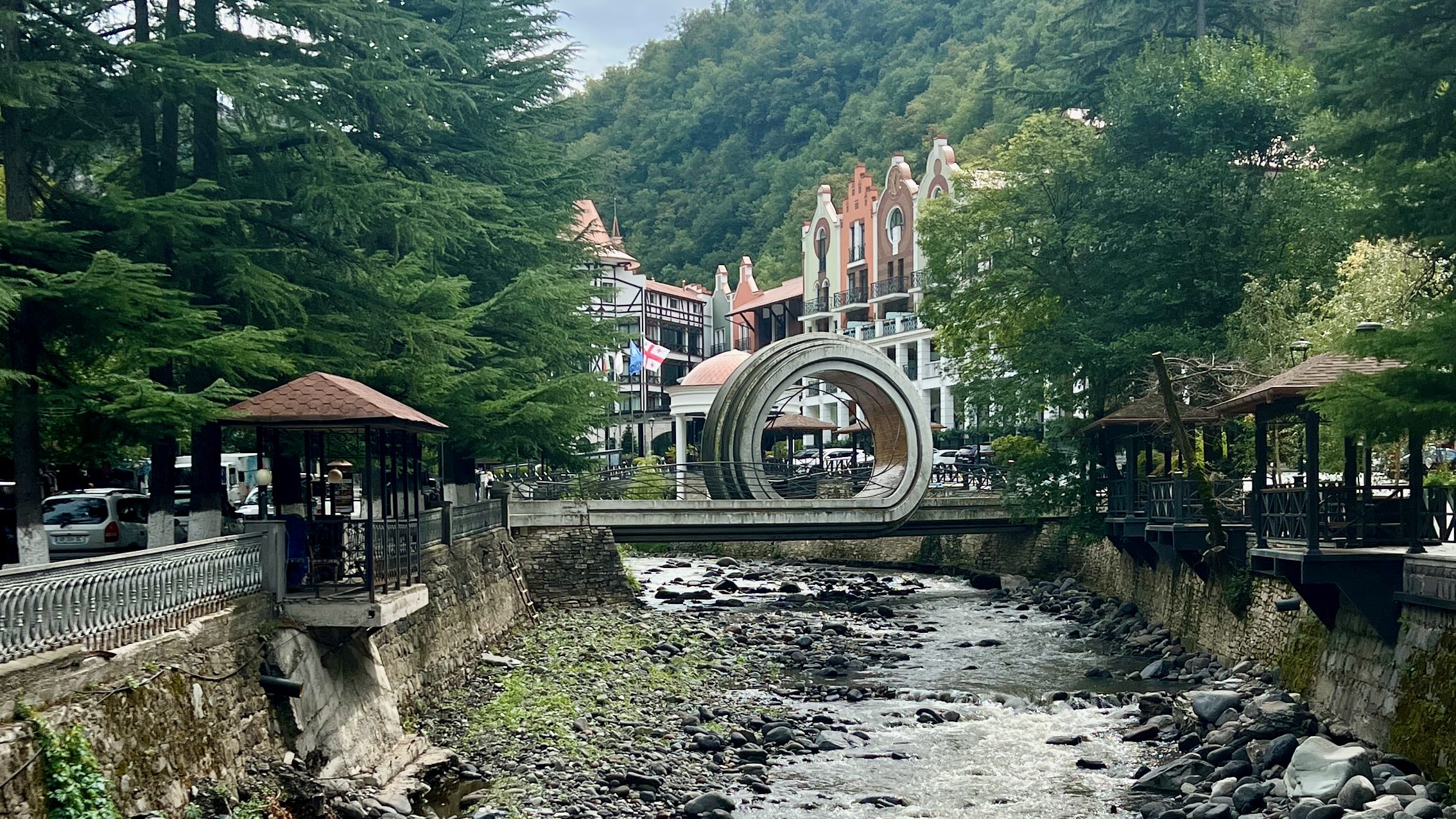 The beauty of a stone bridge with a circular arch spans a rocky stream, surrounded by trees and buildings with red roofs in the mountainous area of Borjomi. This picturesque setting is truly one of its must-see spots and might make you wonder, is Borjomi worth visiting? With such charm, it certainly invites exploration.