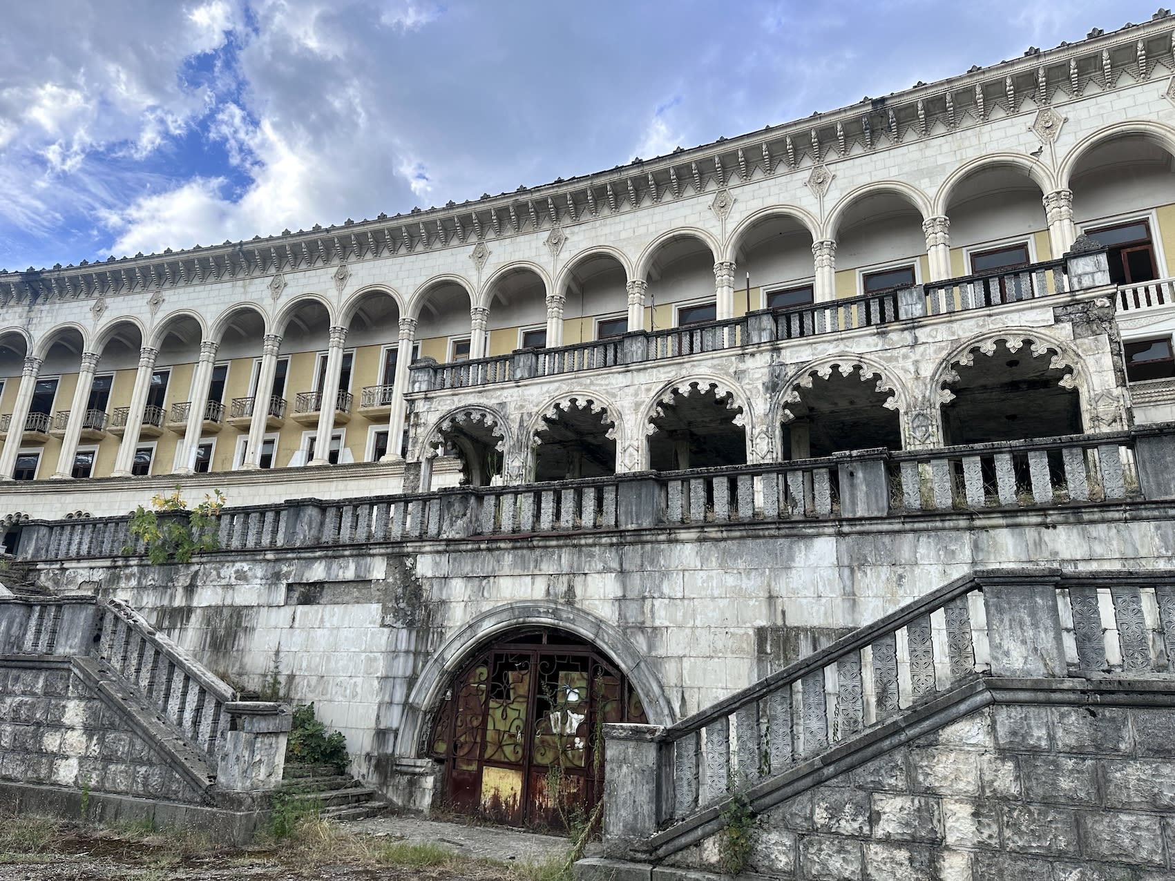 A large, abandoned building with a series of arches and columns on its upper level stands as one of Kutaisi’s cultural gems. The structure has a weathered, stone facade and overgrown staircases leading to a rusted, iron gate. The sky above is partly cloudy, heightening the mystery surrounding this must-see sight.
