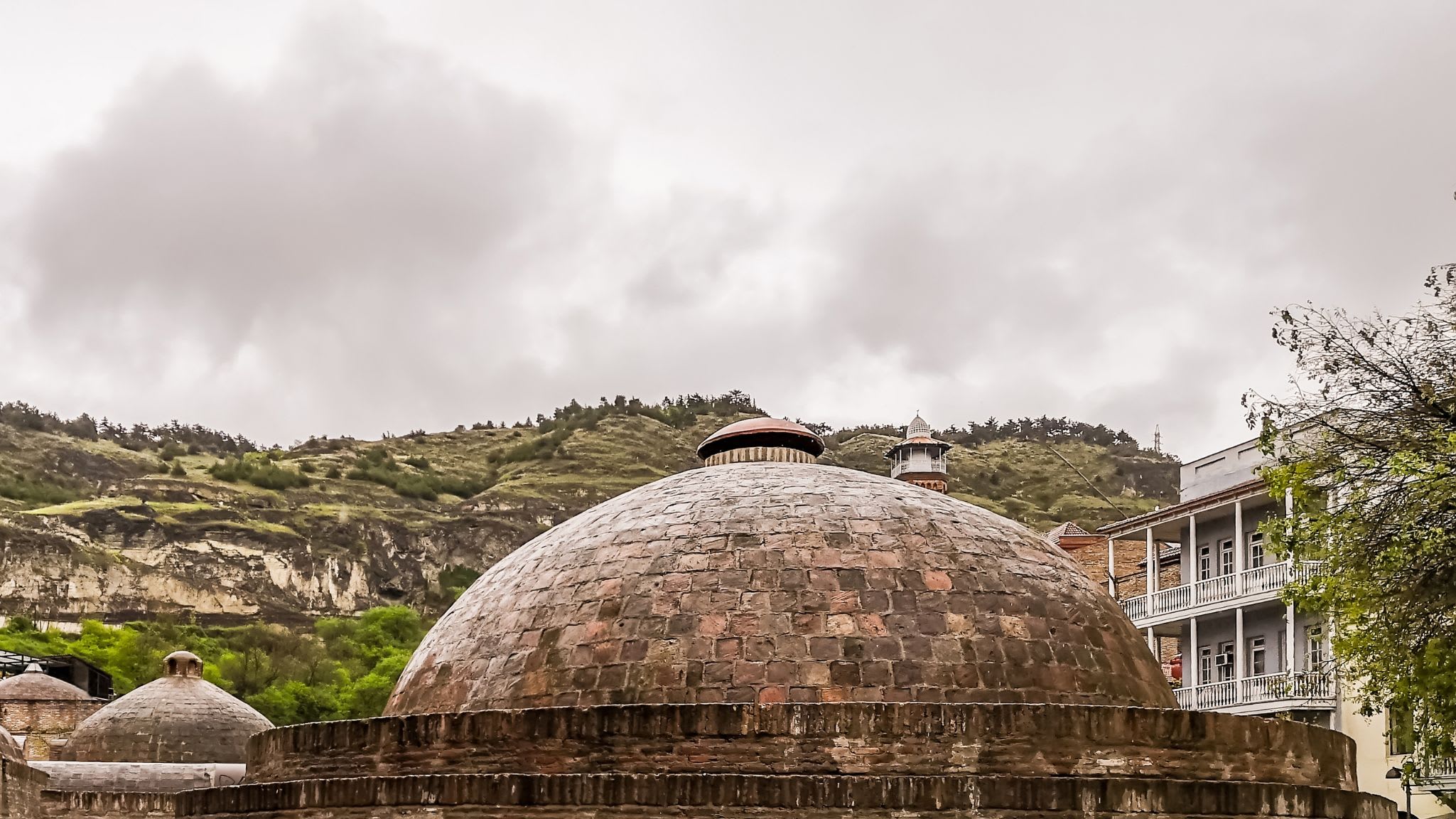 Discover the ancient brick domes of Tbilisi's iconic sulfur baths, nestled among stone buildings with green hills in the background under a cloudy sky. The Tbilisi sulfur baths offer a unique and relaxing experience, inviting visitors to immerse themselves in the warm, healing waters that have been cherished for centuries.