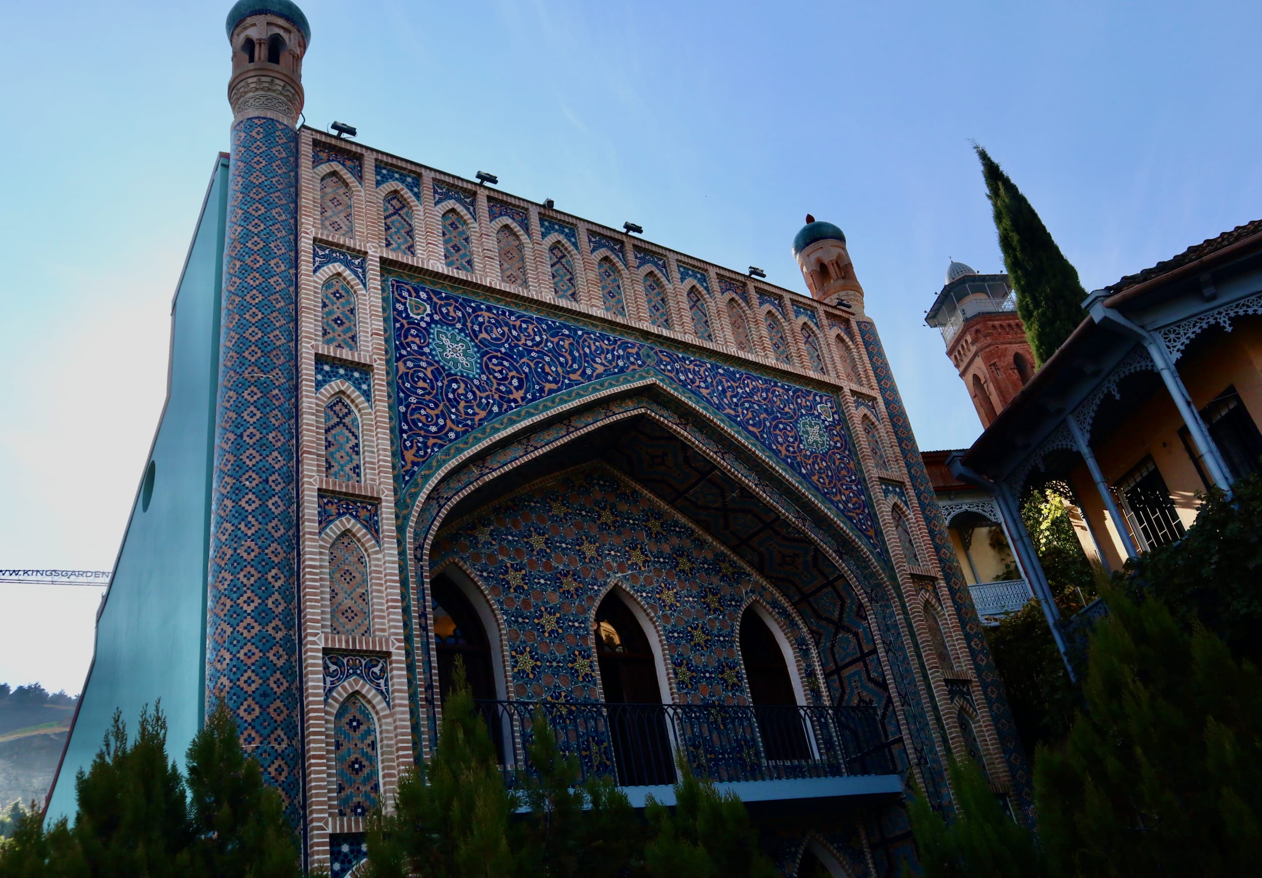 A building with ornate blue and beige tilework, featuring arched windows and two cylindrical towers capped with domes, reminiscent of the architecture in Tbilisi, is a must-visit landmark near the famous Tbilisi sulfur baths.