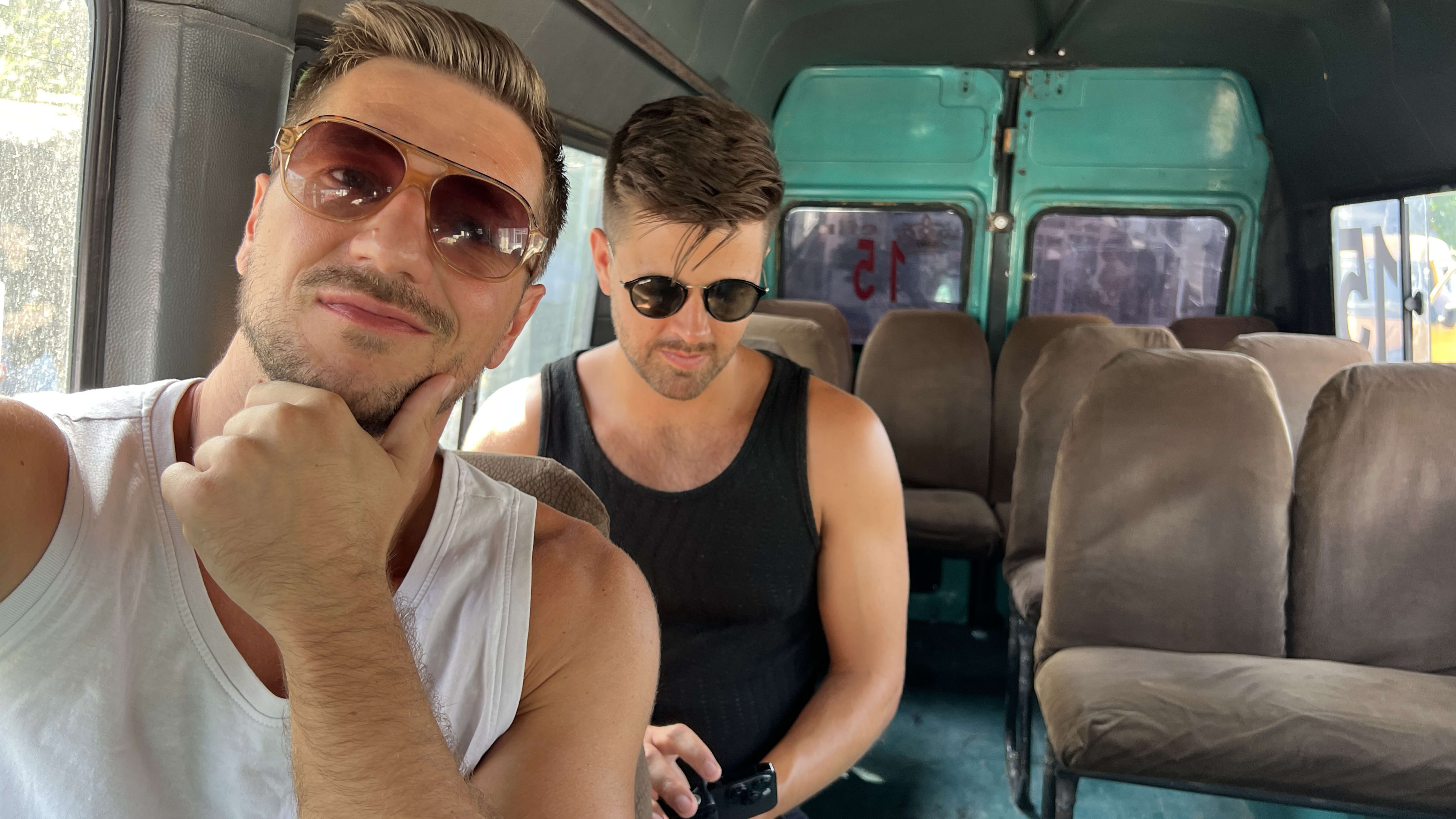 Two men wearing sunglasses sit on a mostly empty bus in Azerbaijan. One, like a thoughtful travel guide, gazes at the camera with a hand on his chin, possibly pondering things to do in Kish Azerbaijan. The other man is deeply engrossed in his device, perhaps researching attractions or planning their next adventure in this scenic region.