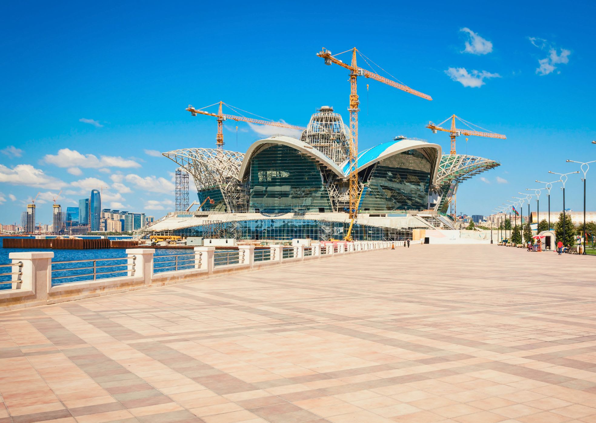 Modern building under construction with large cranes in Baku, showcasing a unique curved design by a waterfront promenade under a clear blue sky. Ideal for those considering if Baku is worth visiting to explore architectural wonders in Azerbaijan's vibrant capital, where innovation meets stunning vistas and cultural richness.