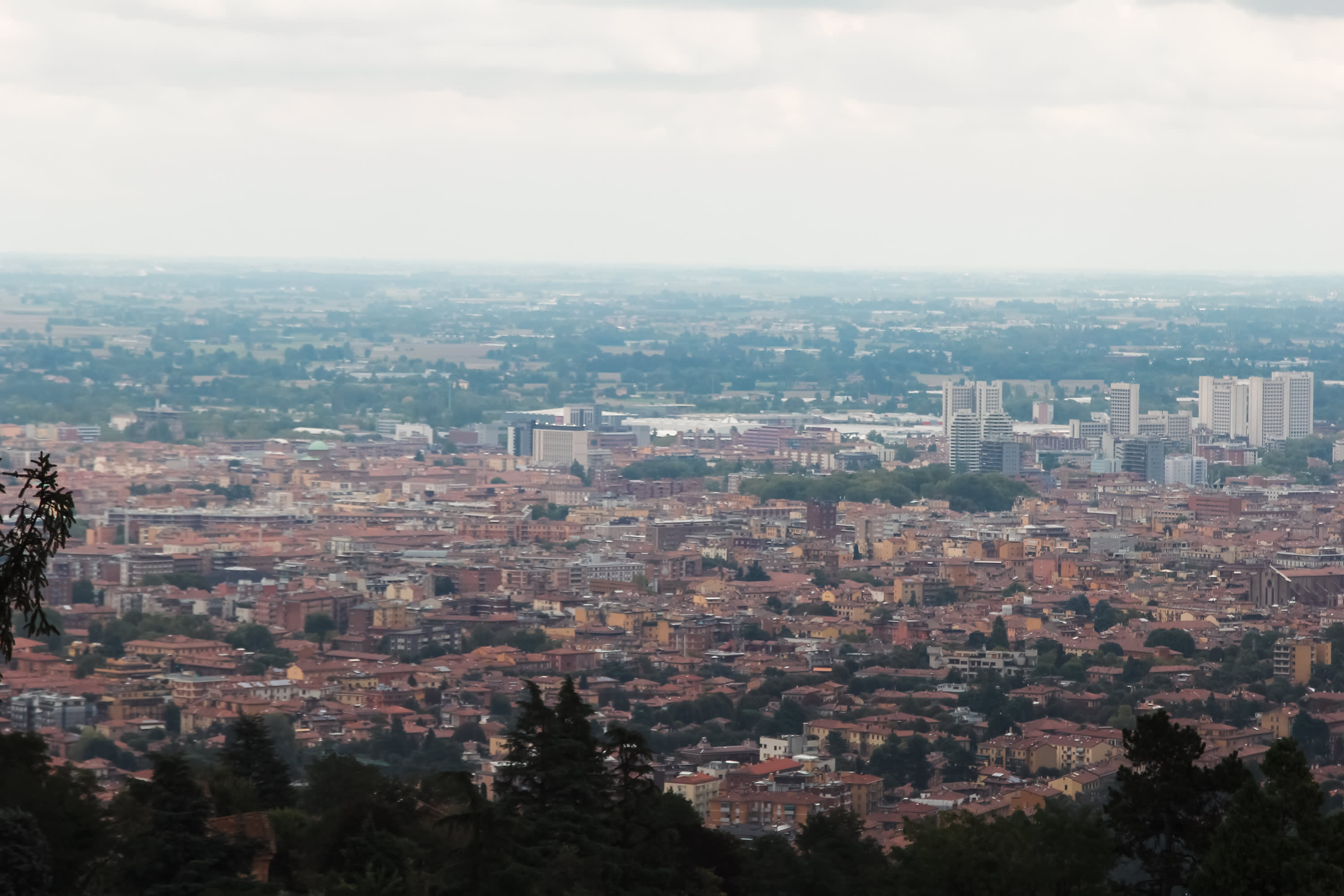 A wide cityscape view shows densely packed red-roofed buildings in the foreground, several tall modern buildings in the background, and a flat landscape extending to the horizon under a cloudy sky.