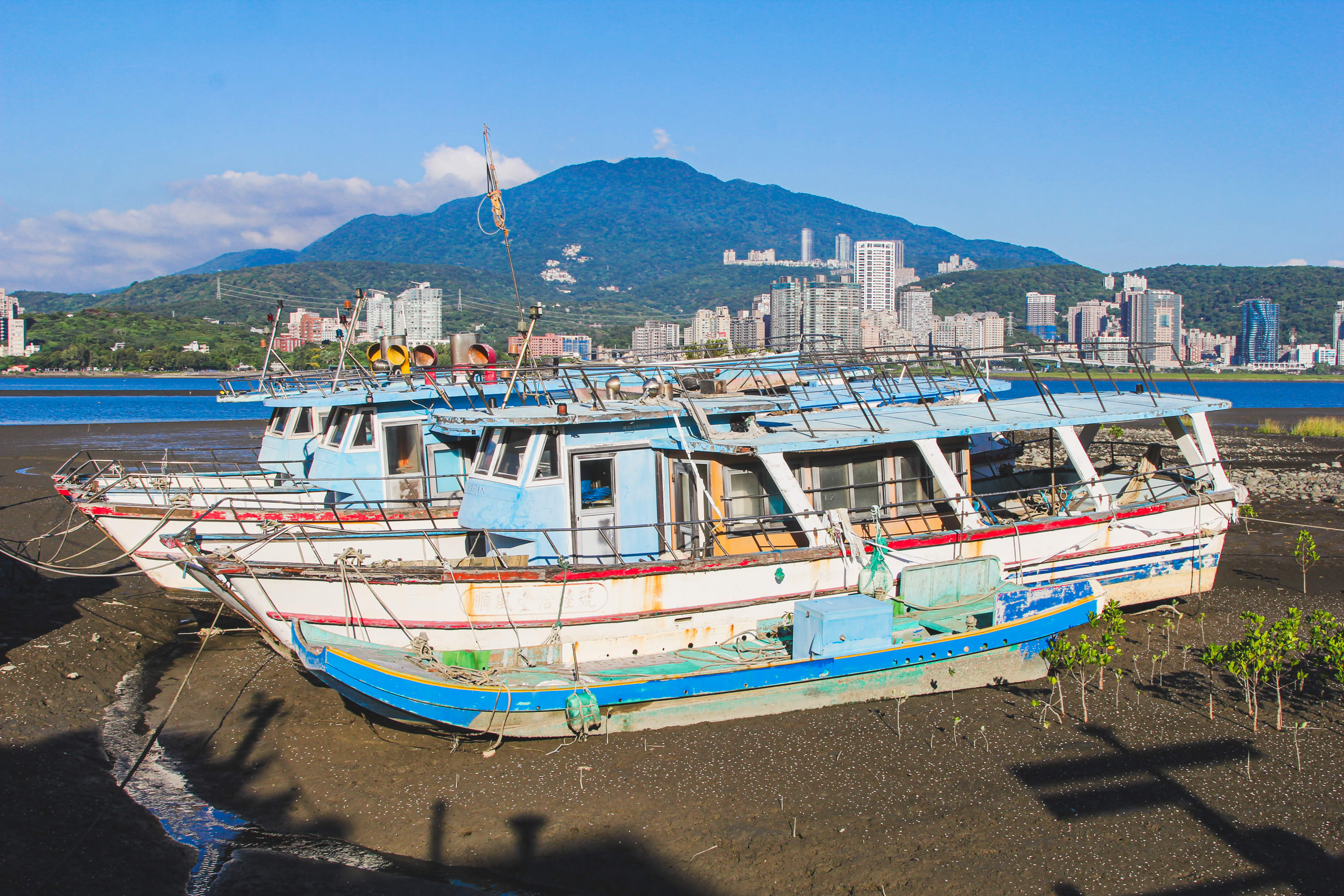Three colorful fishing boats are docked on a muddy shore, with a city skyline, river, and lush green mountains in the background—an inviting scene to discover along popular Taiwan bike routes under a clear blue sky.