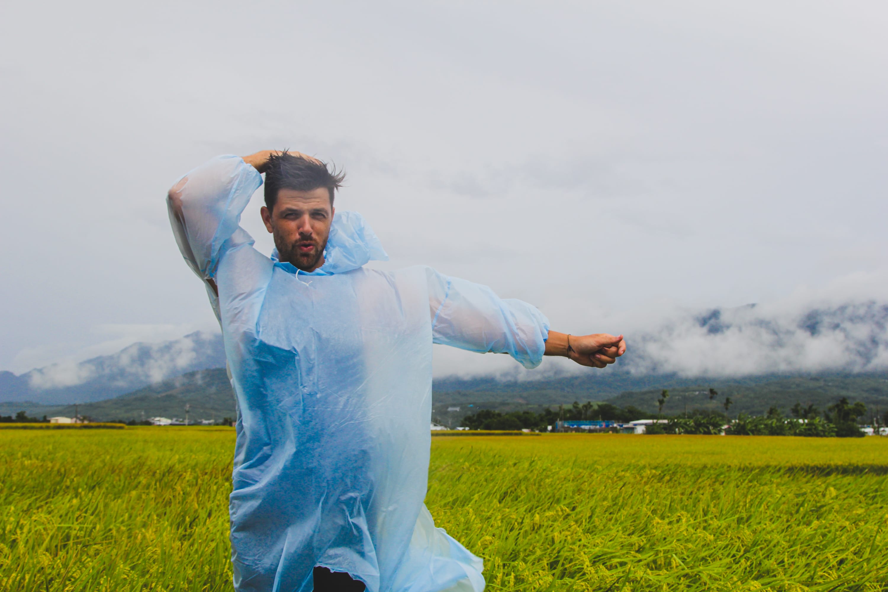 A man wearing a blue rain poncho strikes a playful pose in a green field, with mountains and a cloudy sky behind him—capturing the spirit of exploring scenic cycling routes Taiwan has to offer.