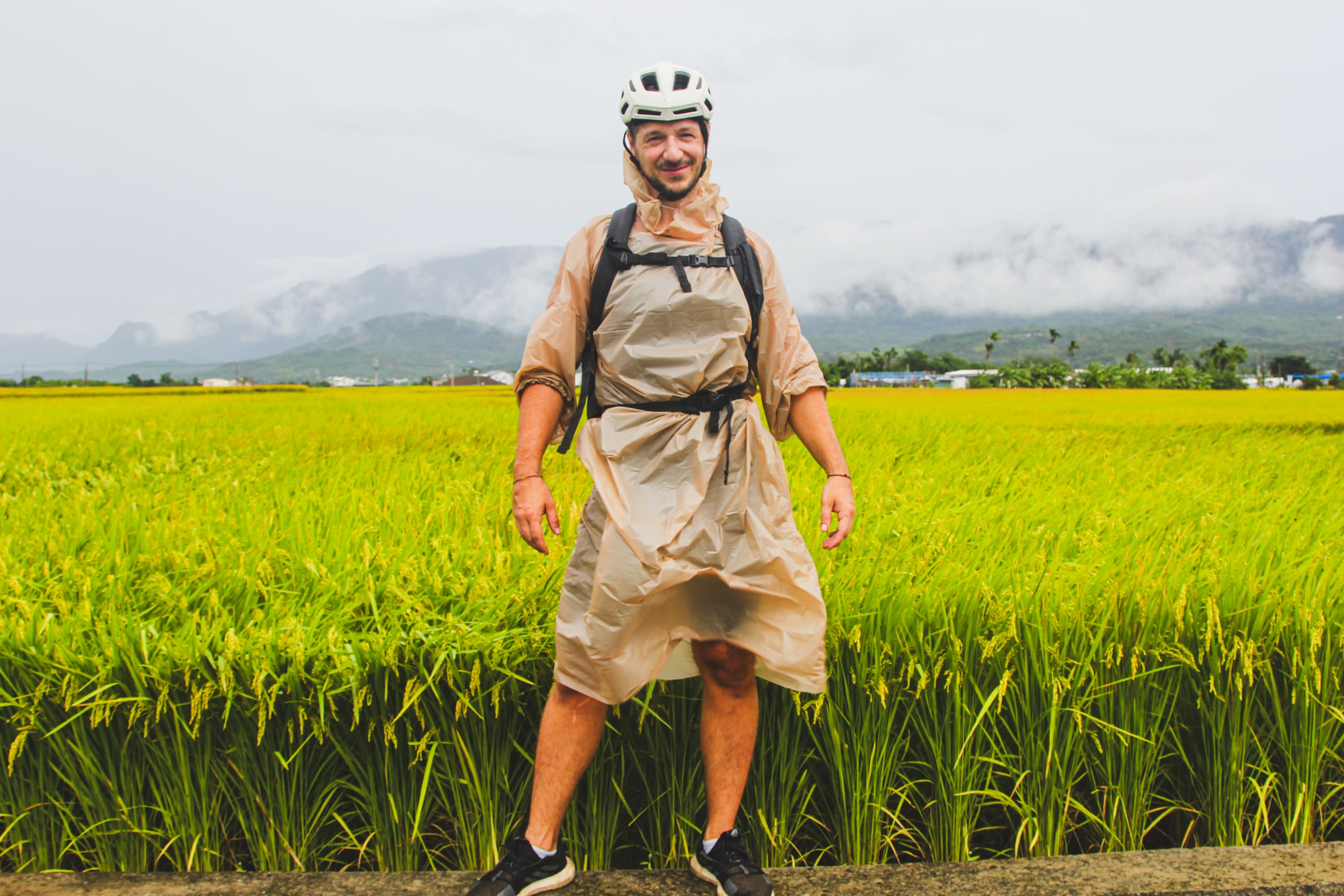 A man wearing a helmet, poncho, and backpack stands smiling in front of a lush green rice field with misty mountains—capturing the spirit of a Taiwan bike adventure.