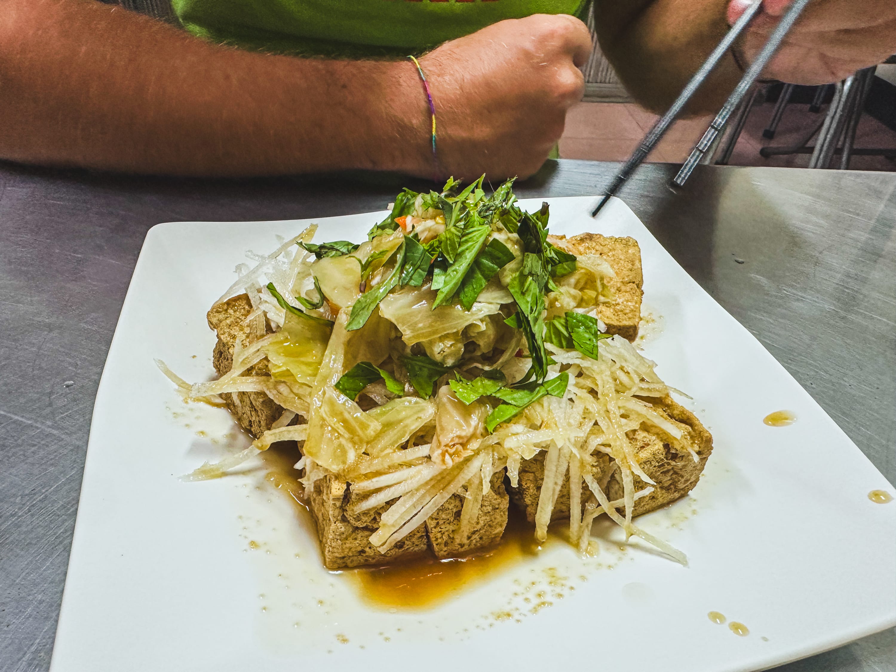 A person uses chopsticks to eat a dish of fried tofu topped with shredded vegetables and herbs, served on a white square plate with sauce—perfect for refueling during a Taiwan bike adventure.