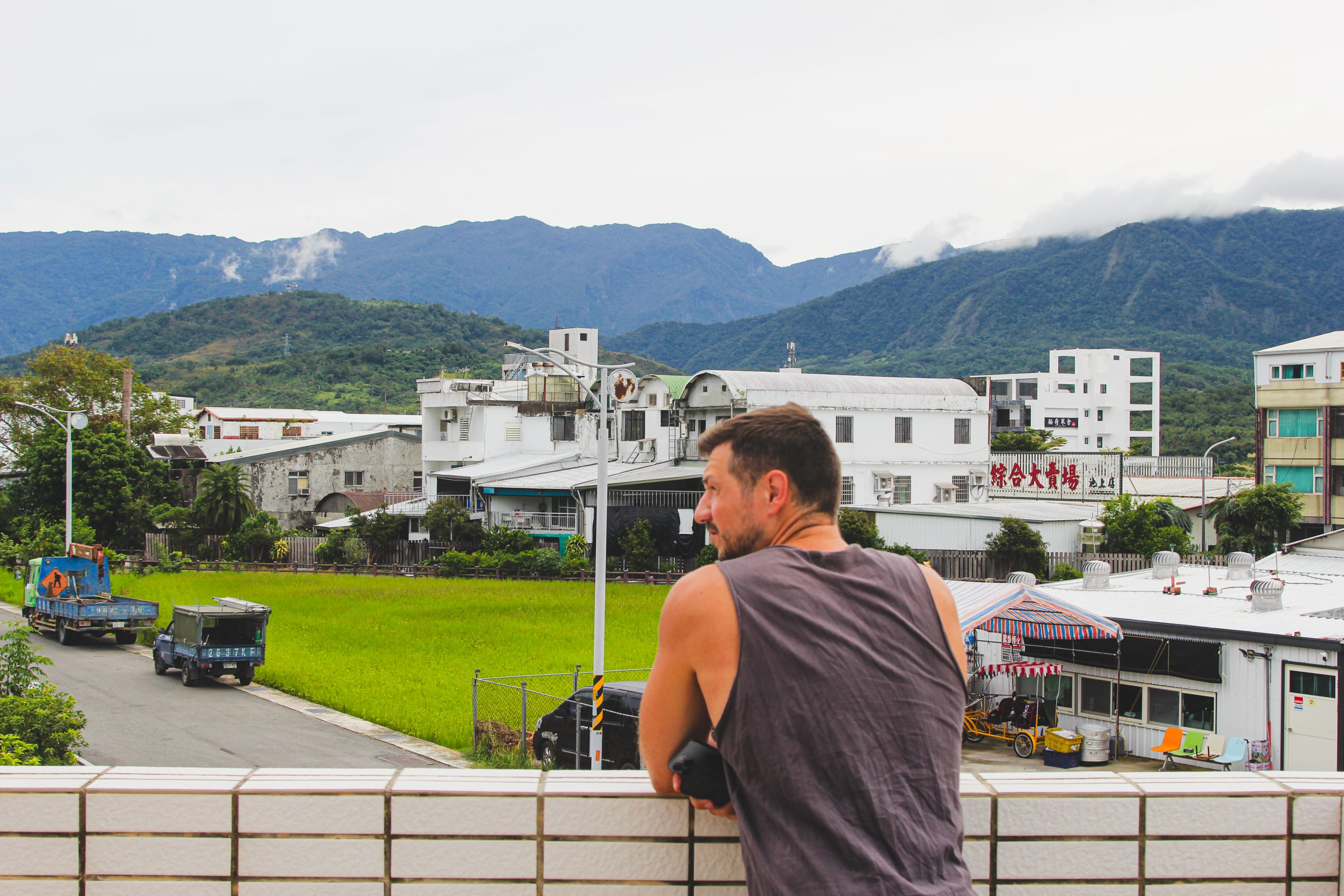 A man in a sleeveless shirt leans on a tiled balcony railing, gazing out over a small town with green fields and mountains—a serene moment for anyone inspired to explore cycling routes Taiwan has to offer.