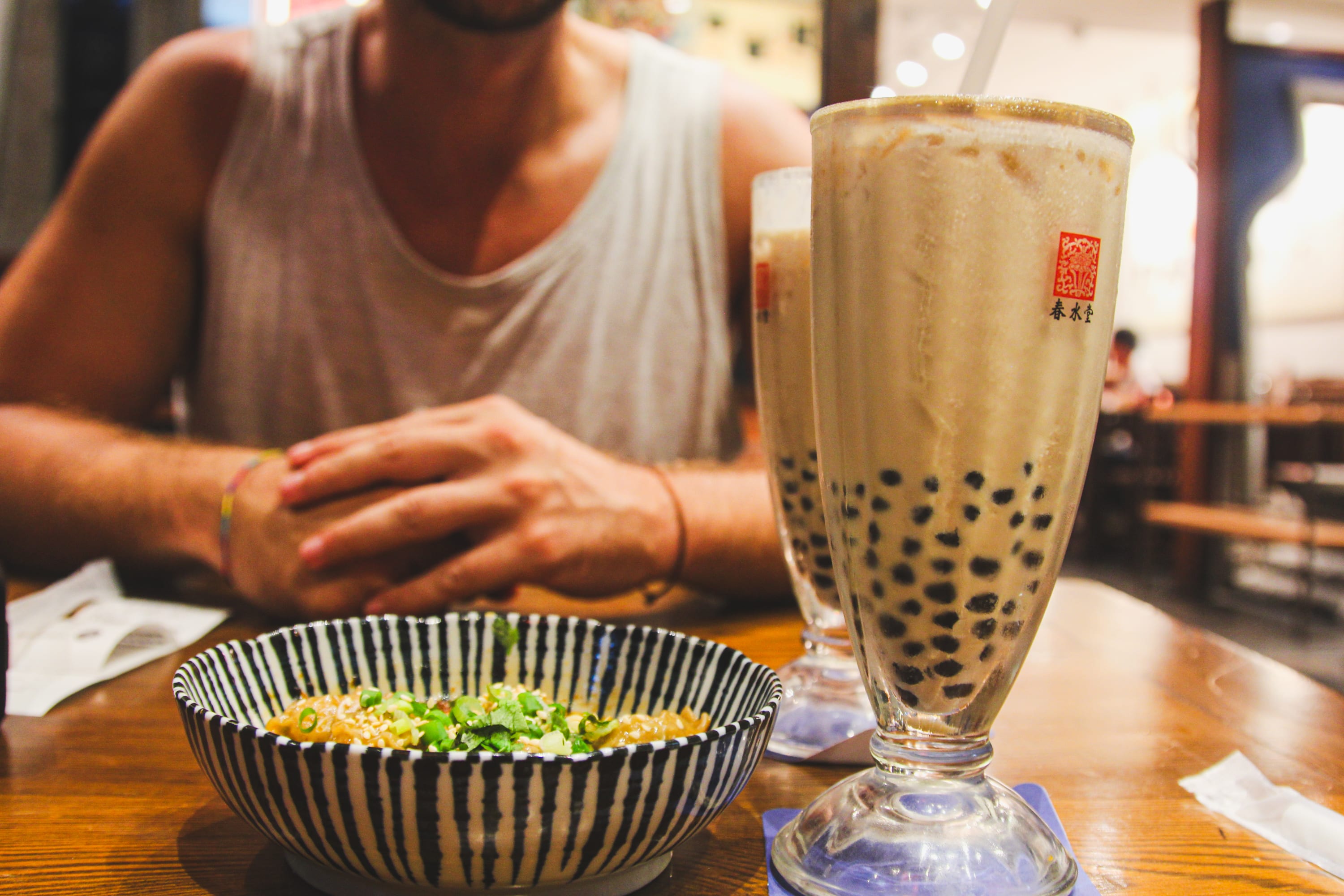 A person in a sleeveless shirt sits at a table with a bowl of noodles topped with green onions and a tall glass of bubble tea—a perfect treat after exploring Taiwan cycling routes. Another glass of bubble tea rests in the background.