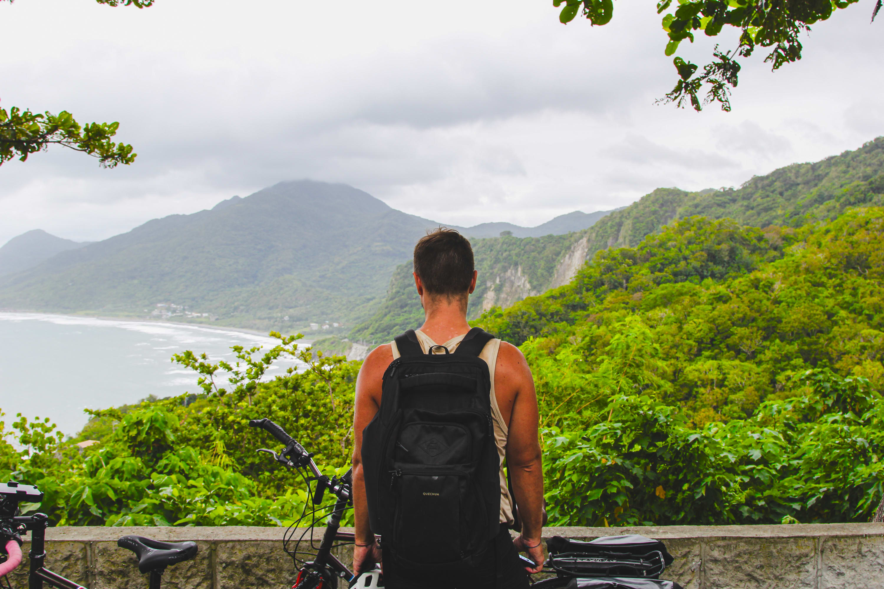 A person with a backpack stands facing lush green mountains and the coast, framed by leafy branches, with a bicycle beside them under a cloudy sky—ready for a cycling adventure along scenic Taiwan bike routes.