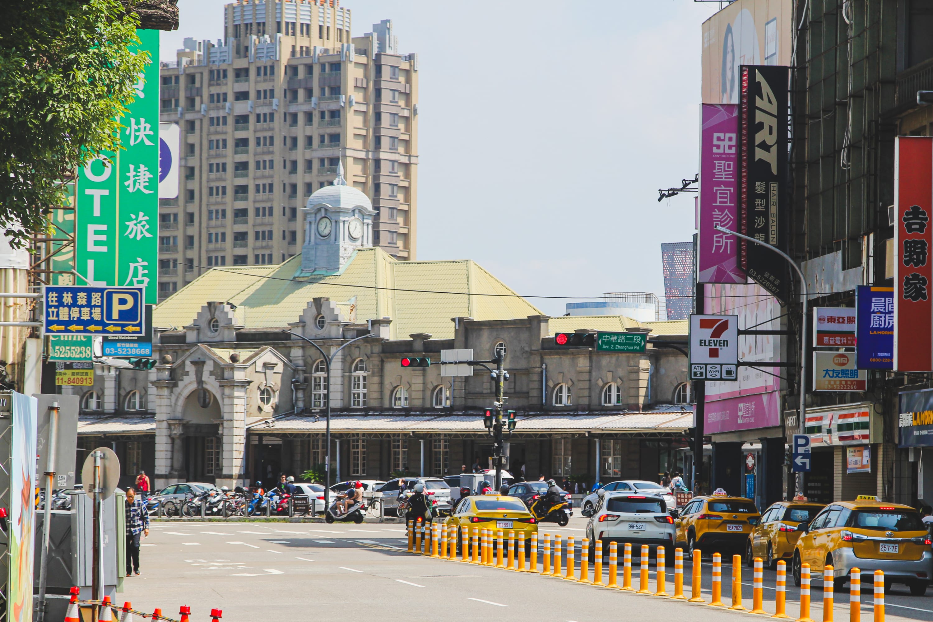 A busy city street with yellow taxis, cars, and scooters near an old stone building with a clock tower. Tall modern buildings and colorful signs in Chinese line the street—a lively stop on many Taiwan cycling routes under a sunny sky.