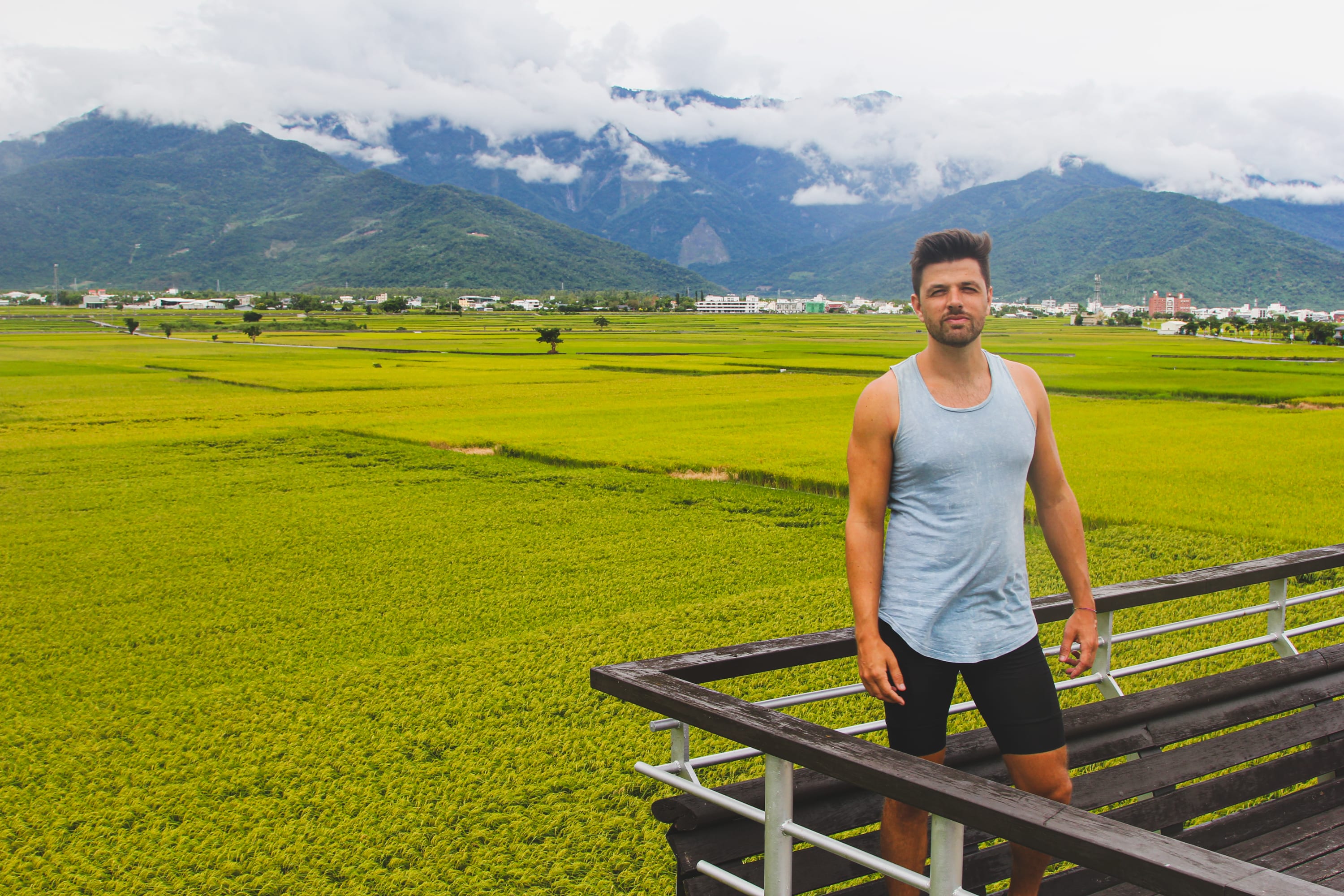 A man in a light gray tank top and black shorts stands on a wooden platform overlooking lush green rice fields—an inspiring stop along scenic Taiwan cycling routes, with mountains and cloudy skies framing the backdrop.