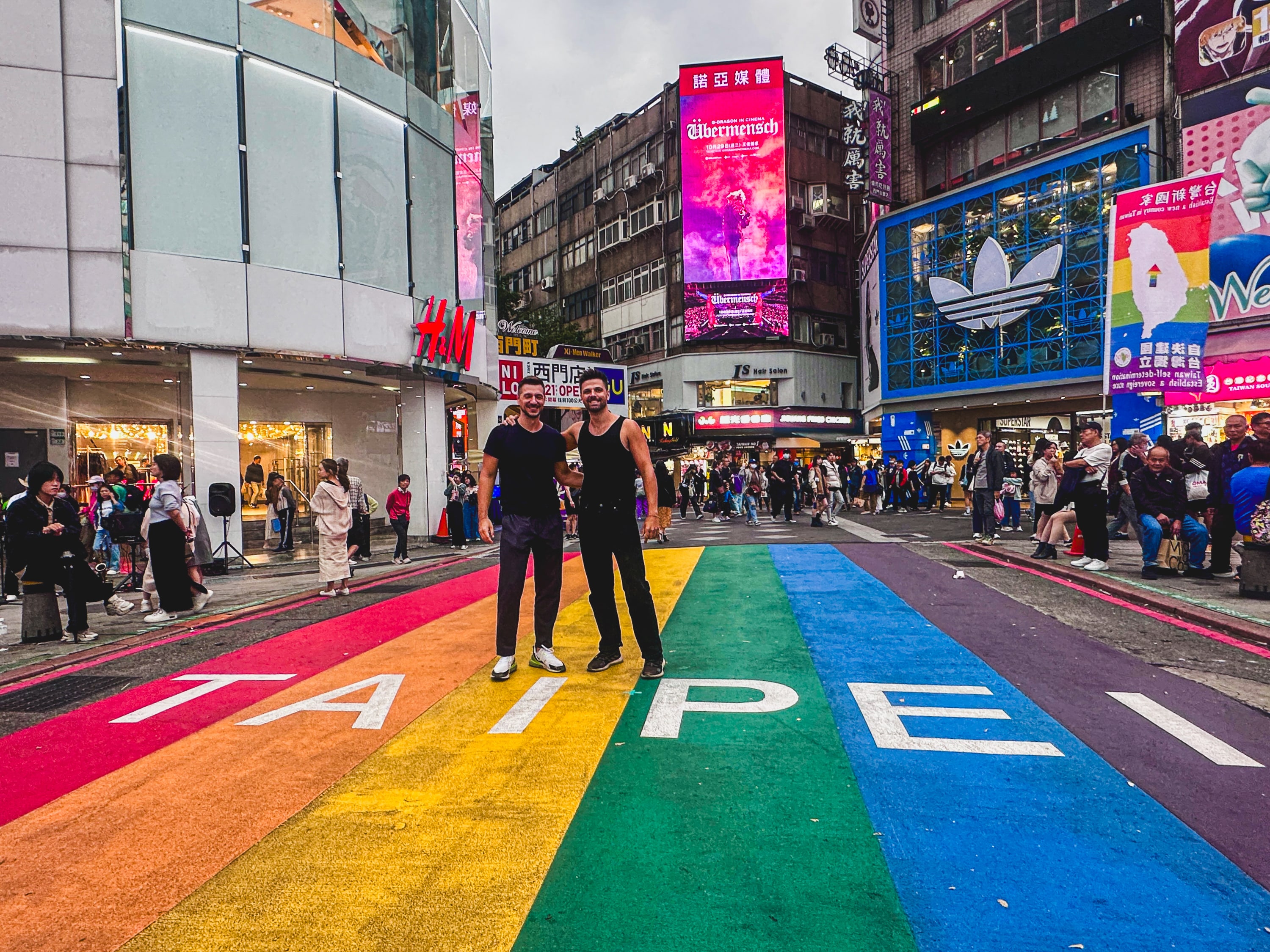 Two people stand on a rainbow-painted crosswalk with 'TAIPEI written on it, surrounded by busy city streets, shops, and colorful billboards—an inviting scene for anyone planning to cycle around Taiwan or explore famous Taiwan cycling routes.