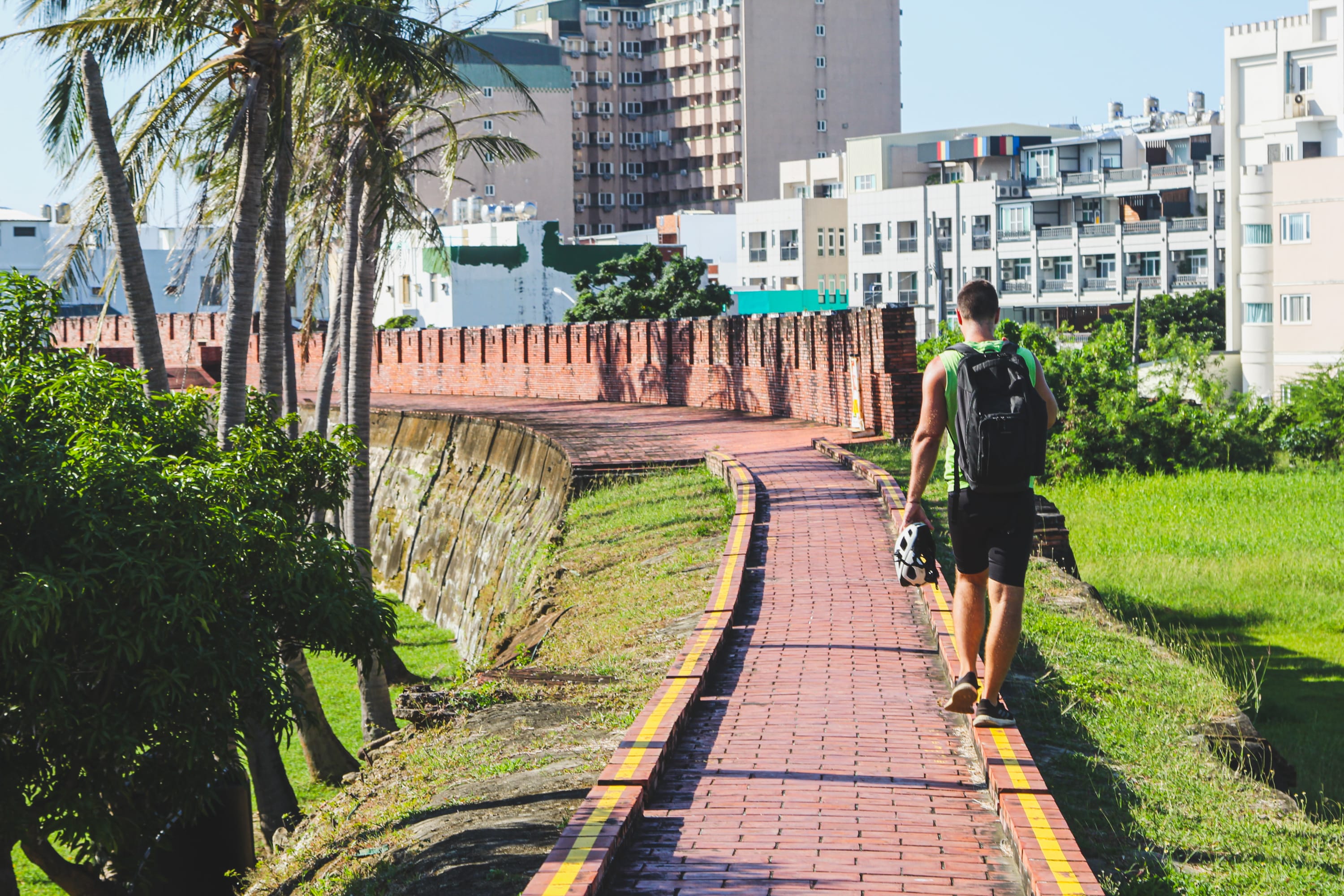 A man with a backpack walks along a red brick path beside an ancient city wall, palm trees, and green grass—an inviting scene familiar to those exploring Taiwan cycling routes under a clear blue sky.