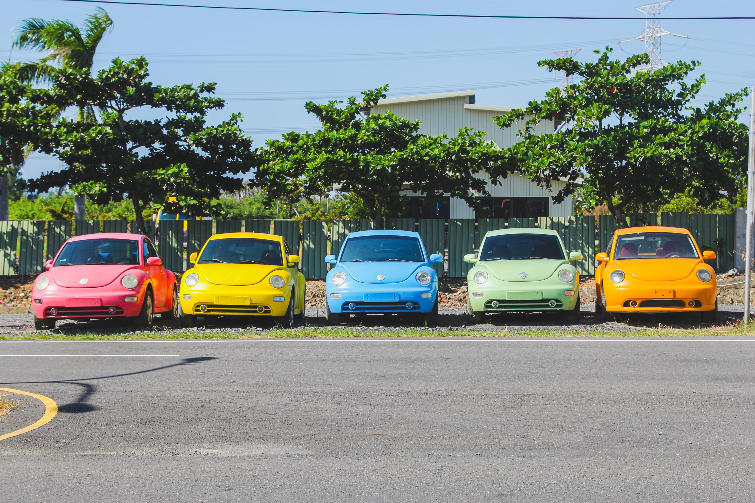 Five Volkswagen Beetle cars in vibrant colors are parked in a row on gravel near a road, inviting passersby to pause before continuing their Taiwan bike adventure or exploring popular Taiwan cycling routes on a sunny day.