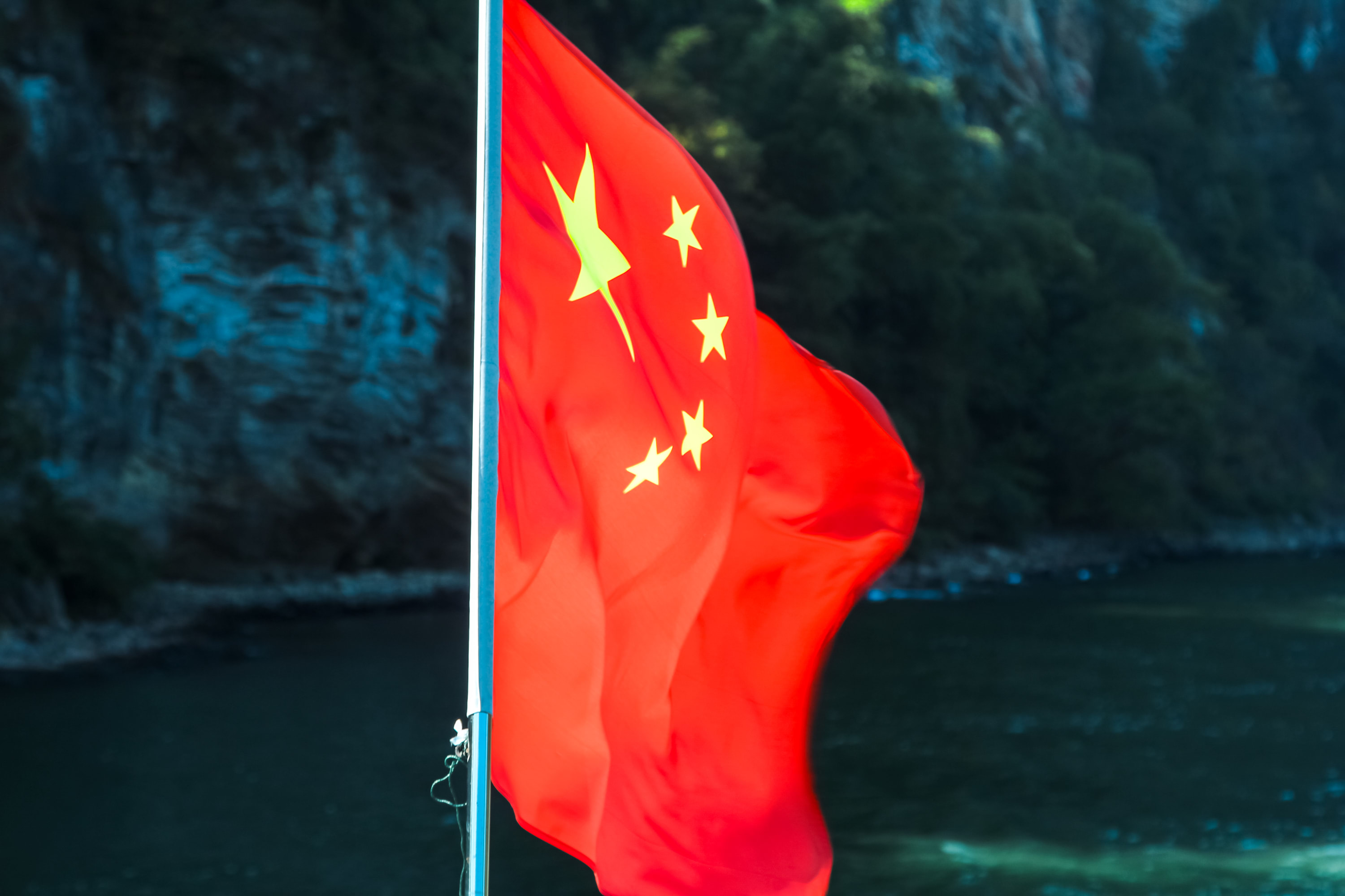 The image shows the national flag of China, featuring a large yellow star and four smaller stars on a red background, waving outdoors near a body of water along the scenic Guilin to Yangshuo cruise route, with trees and rocky cliffs in the background.