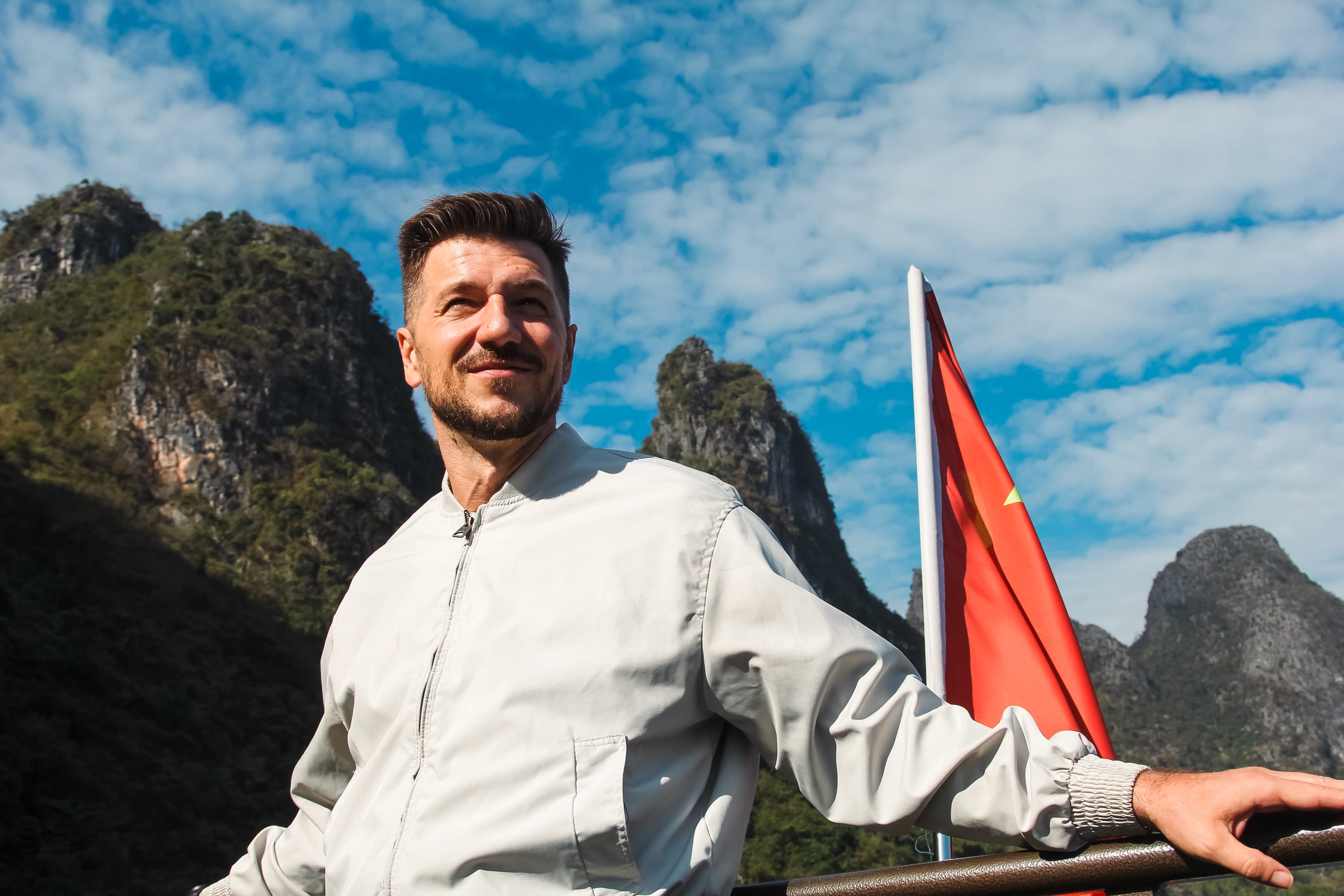 A man in a light jacket stands outdoors, smiling with mountains and a partly cloudy sky behind him. A red flag with yellow stars, likely the flag of China, suggests he may be on a Guilin to Yangshuo cruise adventure.