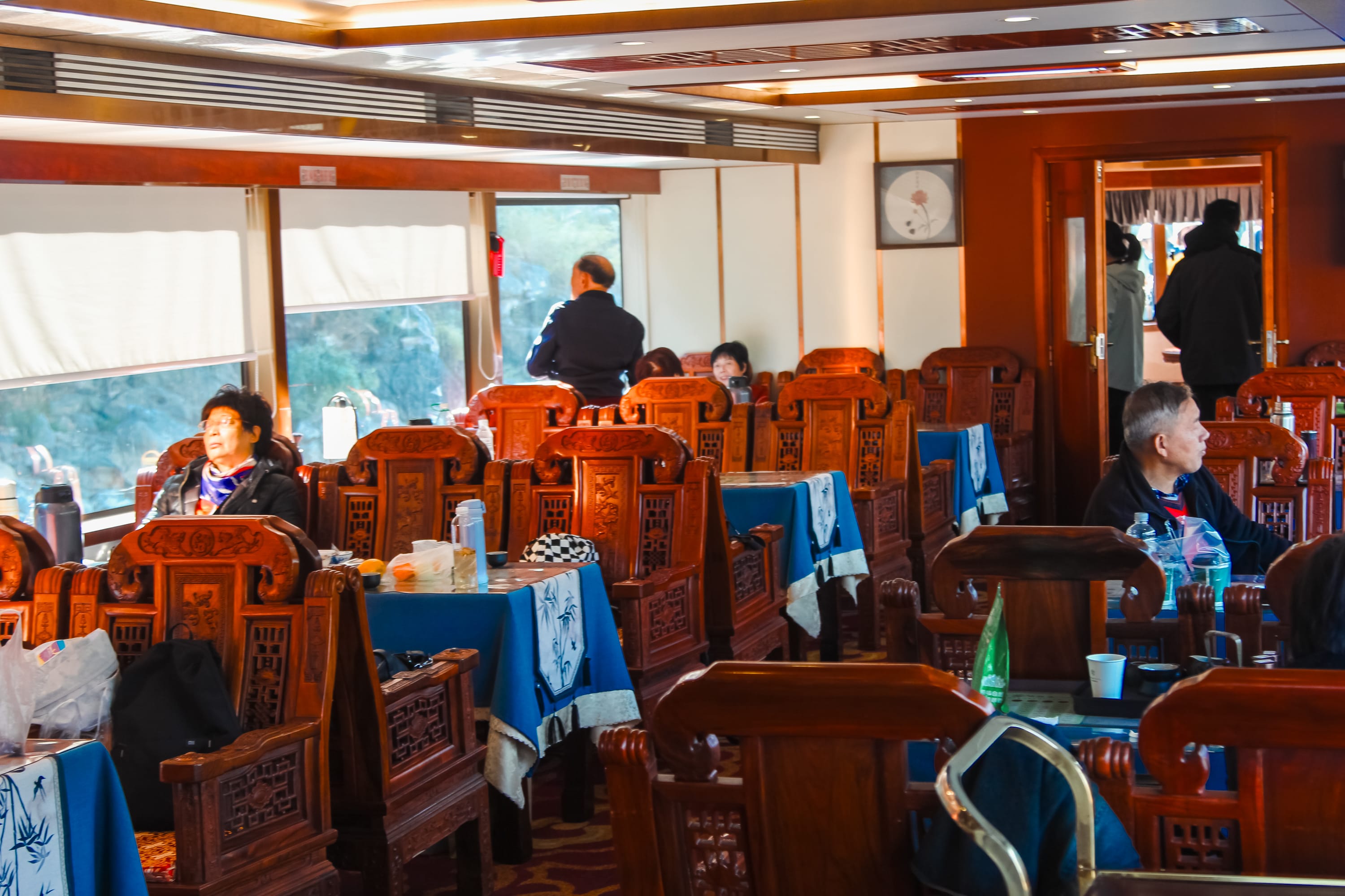 People sit and relax at wooden tables and chairs in a brightly lit restaurant with blue tablecloths, sunlight streaming through large windows—an inviting spot to discuss travel or consult a Yangshuo cruise guide before you book cruise tickets for your Guilin to Yangshuo adventure.