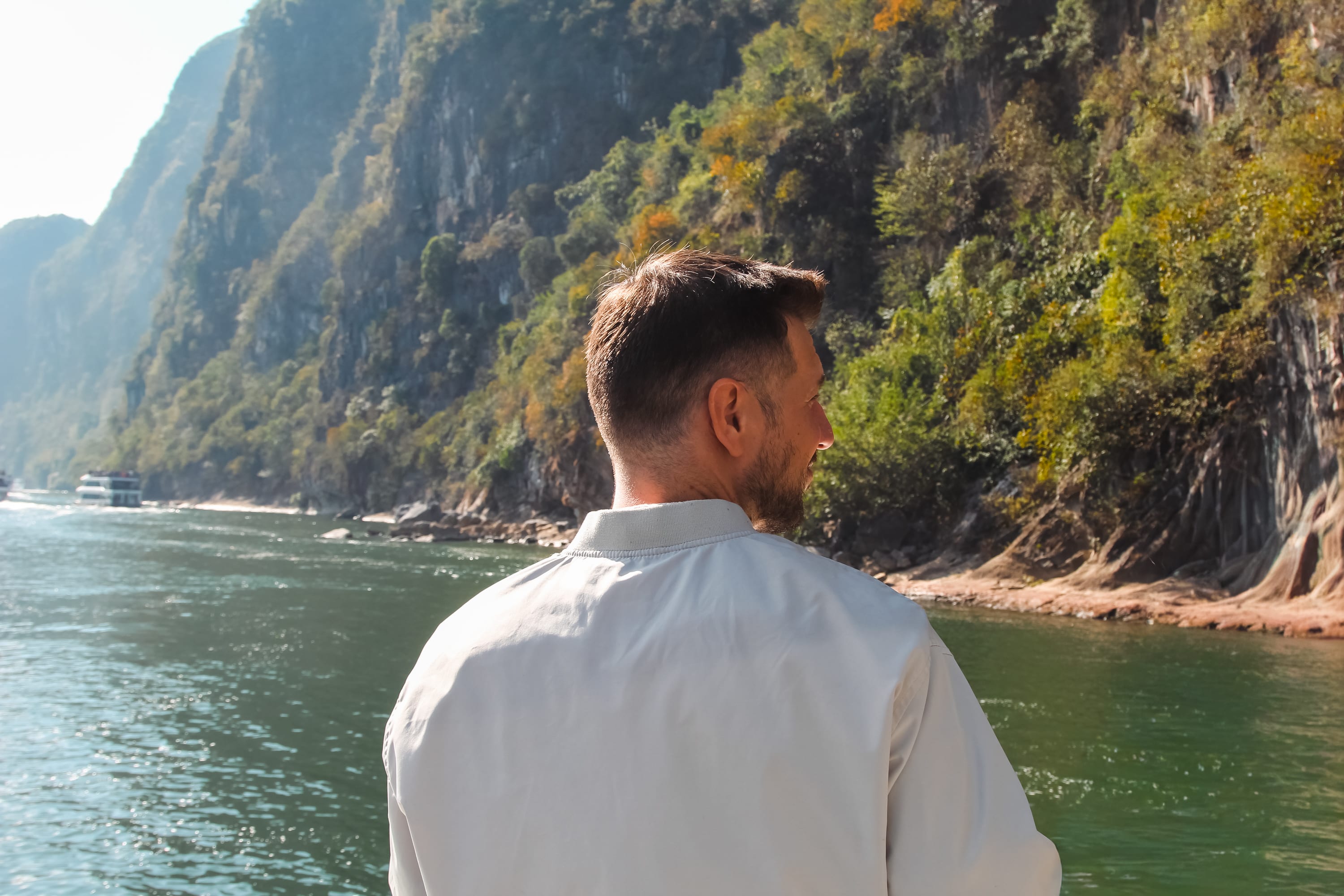 A man in a white jacket stands with his back to the camera, overlooking the river's scenic beauty—a view often admired on a Guilin to Yangshuo cruise—framed by lush green mountains and rocky cliffs under sunlight.