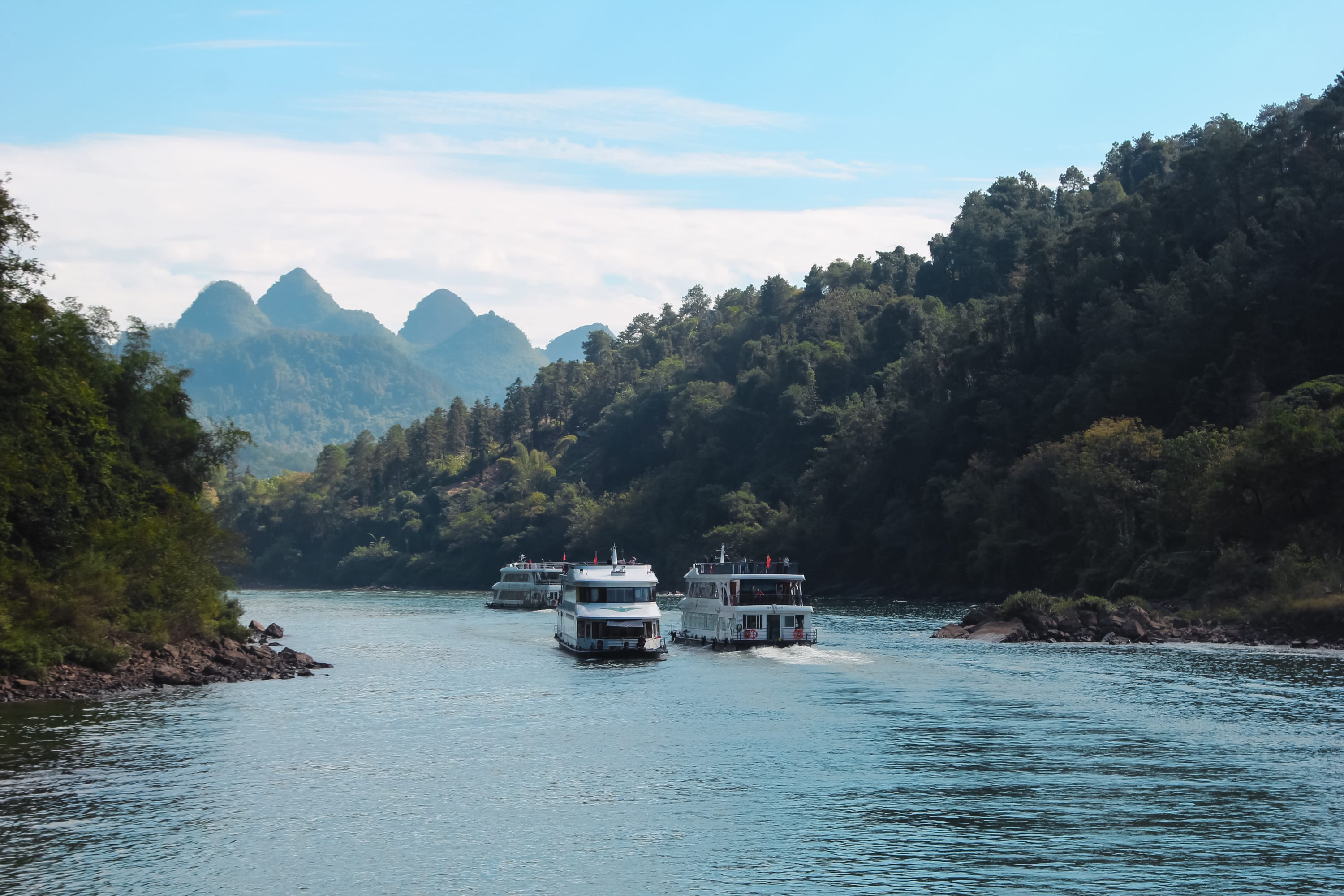 Several white boats travel along a calm river surrounded by lush green hills and distant pointed mountains under a blue sky with wispy clouds, offering a picturesque scene perfect for any Guilin to Yangshuo cruise adventure.