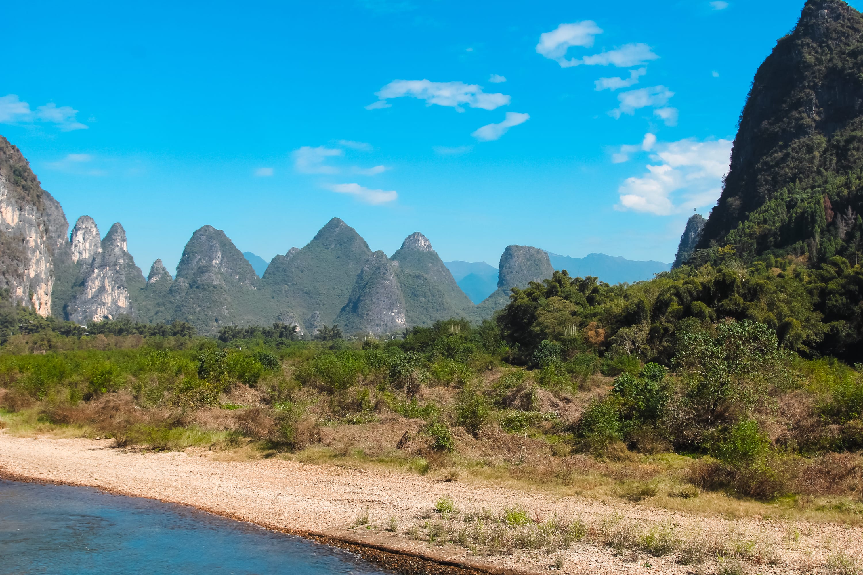 Riverbank with pebbles in the foreground, lush green vegetation in the middle, and dramatic karst mountains under a bright blue sky—the stunning scenery you’ll enjoy on a Guilin to Yangshuo cruise. Book tickets for an unforgettable journey.
