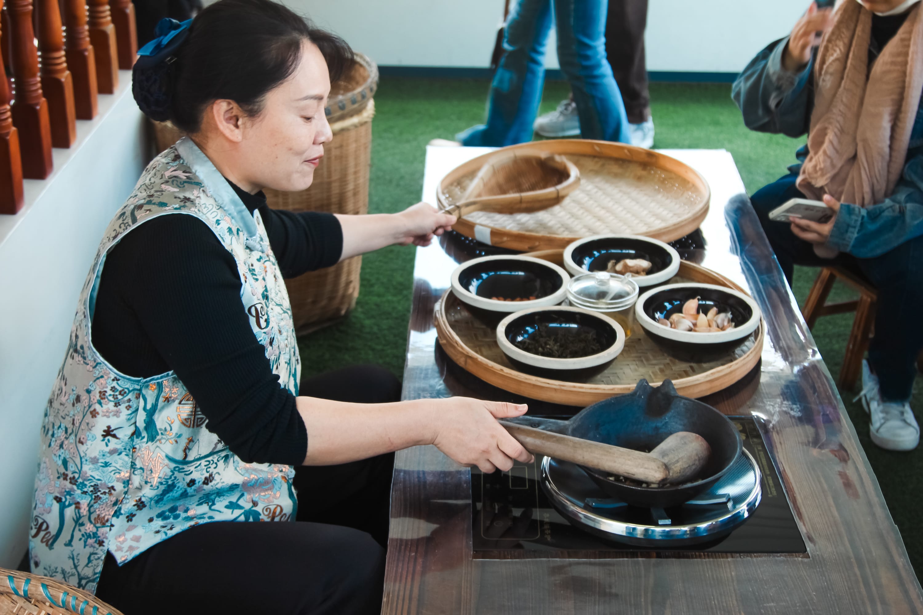 A woman sits at a table, stirring food in a black pan over a burner. Several bowls with various ingredients and a large woven tray are on the table. Other people, like those following a cruise guide, stand nearby, some holding phones.