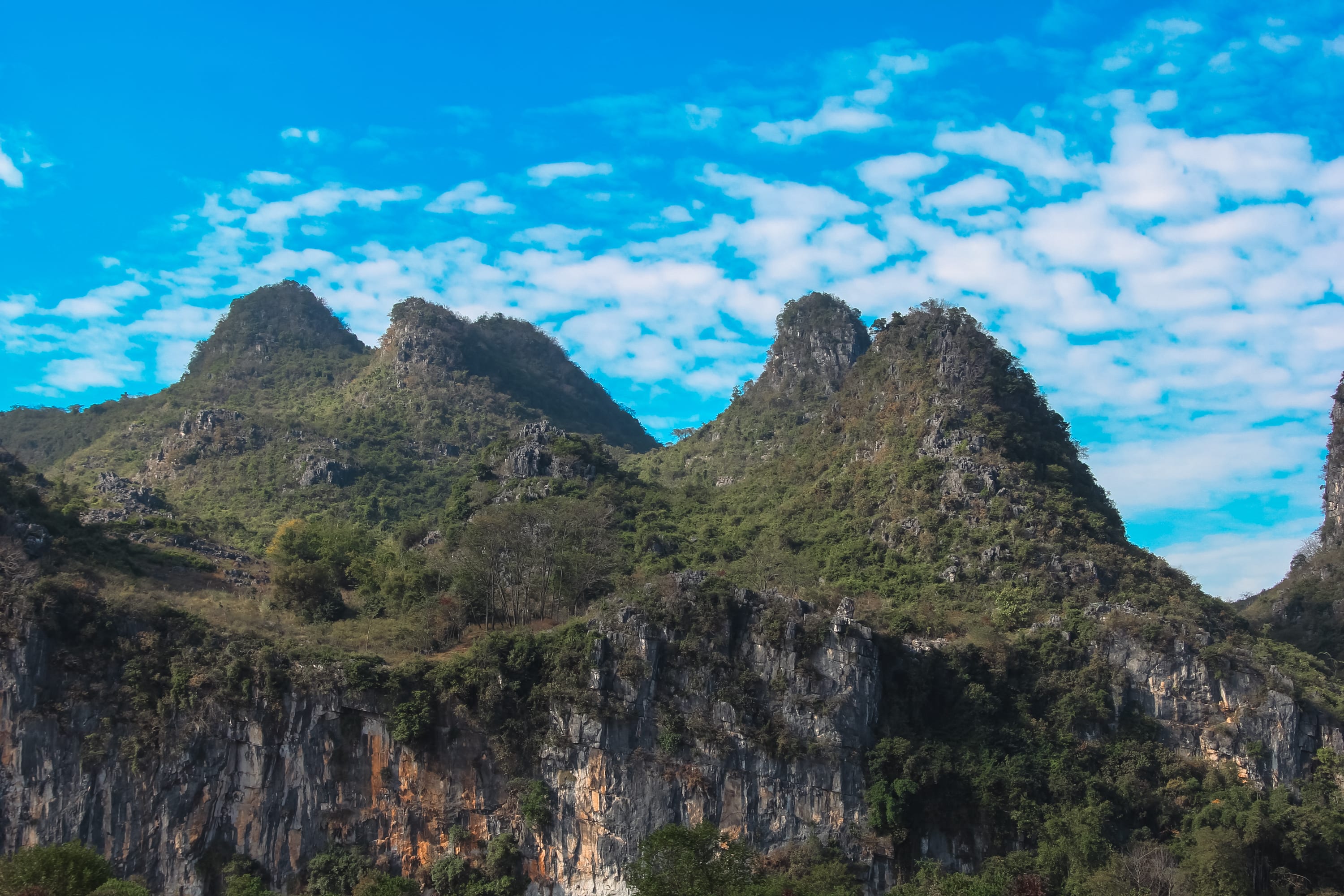 Green, rocky mountains under a bright blue sky with scattered white clouds; the foreground features cliffs with visible rock faces and patches of vegetation—perfect scenery for your Guilin to Yangshuo cruise adventure.