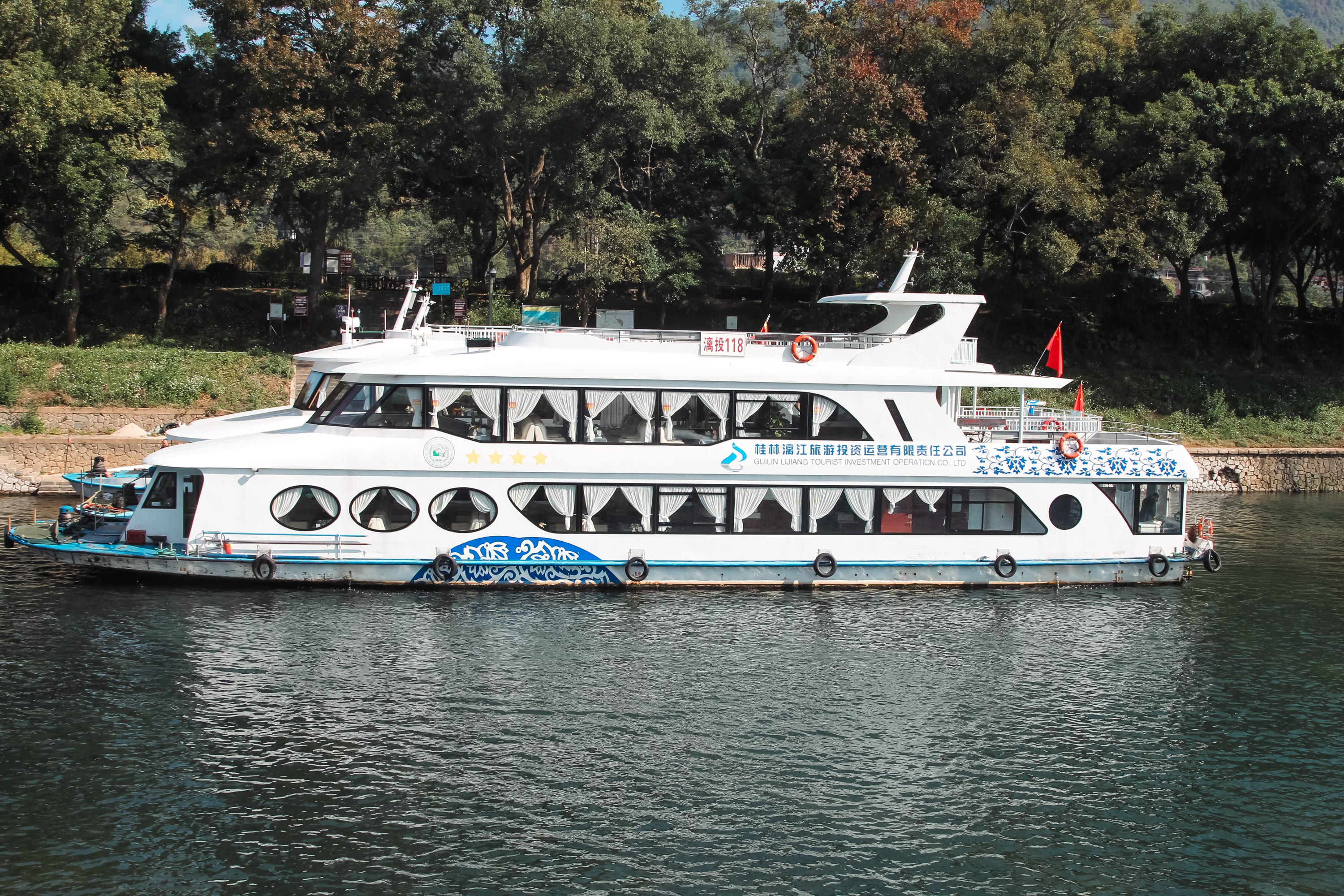 A white and blue river cruise boat with large windows and a red flag sails near a green, tree-lined shore under clear skies, perfect for those following the Yangshuo cruise guide or planning to book cruise tickets from Guilin to Yangshuo.