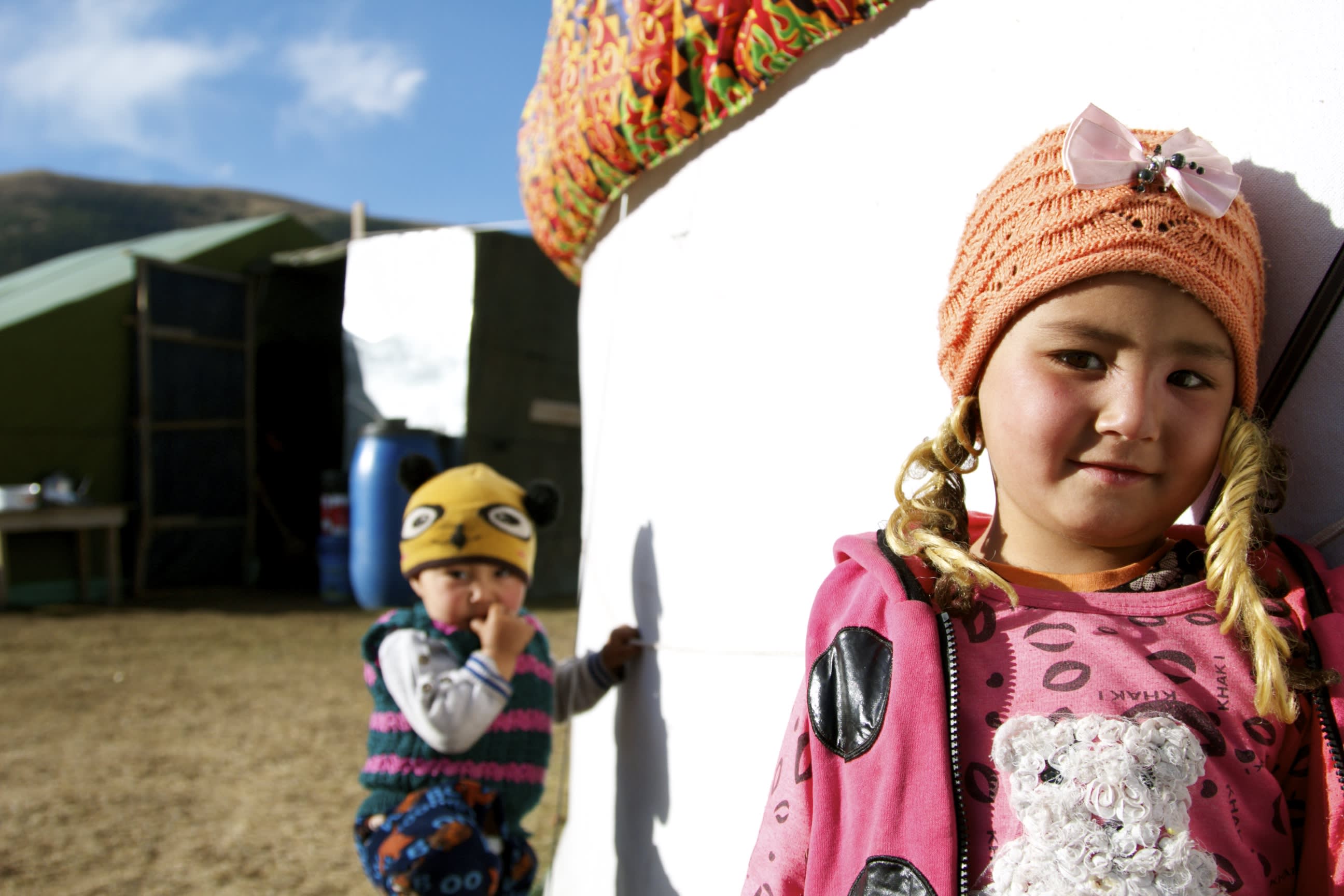 A young girl wearing an orange knit hat with braids stands in front of a yurt, smiling slightly. A younger child in a yellow animal hat and colorful clothing stands in the background next to other tents under the stunning landscapes of Xinjiang's clear blue sky.
