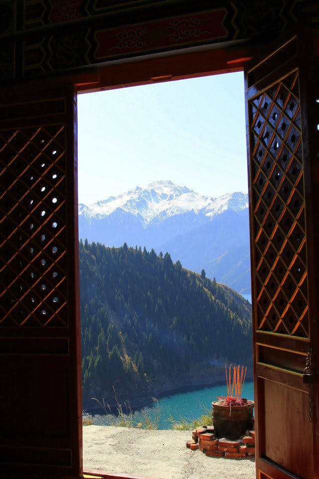 A scenic view through an open doorway reveals the snow-capped mountains of Xinjiang and a forested hillside surrounding a turquoise lake. The foreground features incense sticks in a holder, adding a touch of Uyghur culture to the stunning, tranquil landscape.