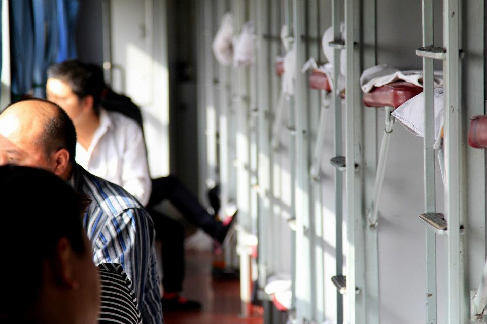 Passengers sit in a train compartment with folded beds and clothes visible, as sunlight streams through the windows, casting shadows. The focus is on the interior setting, offering a glimpse into Xinjiang's stunning landscapes beyond.