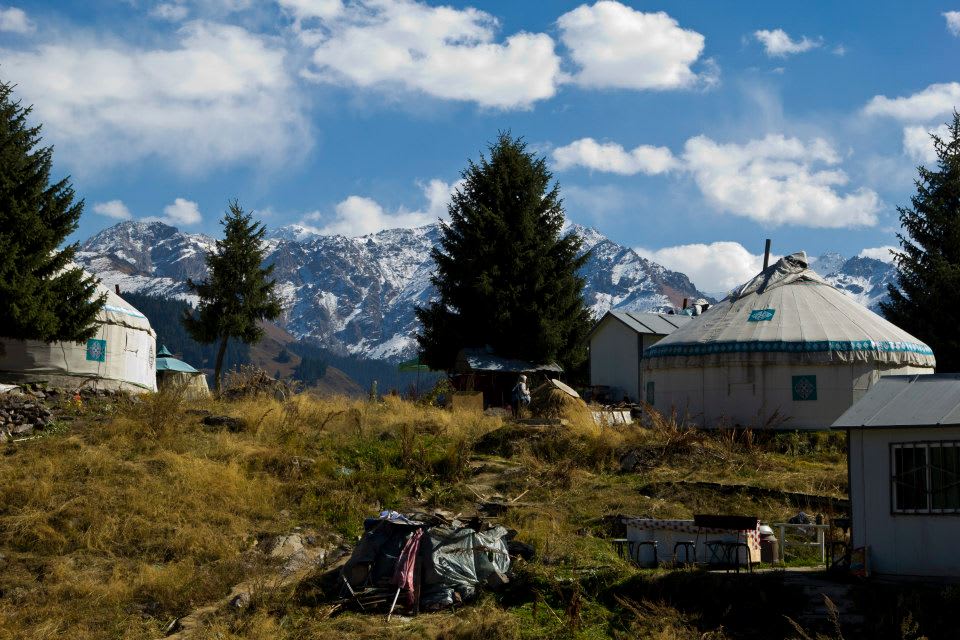 Yurts in a hilly landscape with pine trees, set against a backdrop of stunning, snow-capped mountains and a partly cloudy sky, evoke the rich heritage of Uyghur culture found in Xinjiang.