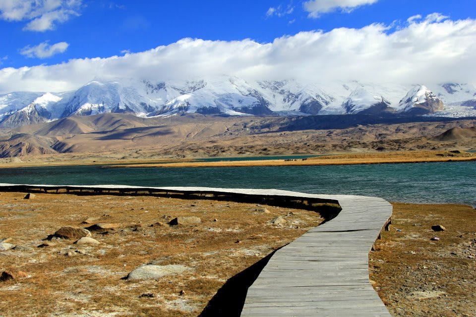 A wooden boardwalk meanders across the dry, rocky terrain of Xinjiang, guiding visitors toward a turquoise lake. Snow-capped mountains rise in the background under a blue sky with scattered clouds, creating a tapestry of stunning landscapes reminiscent of Uyghur culture's rich heritage.