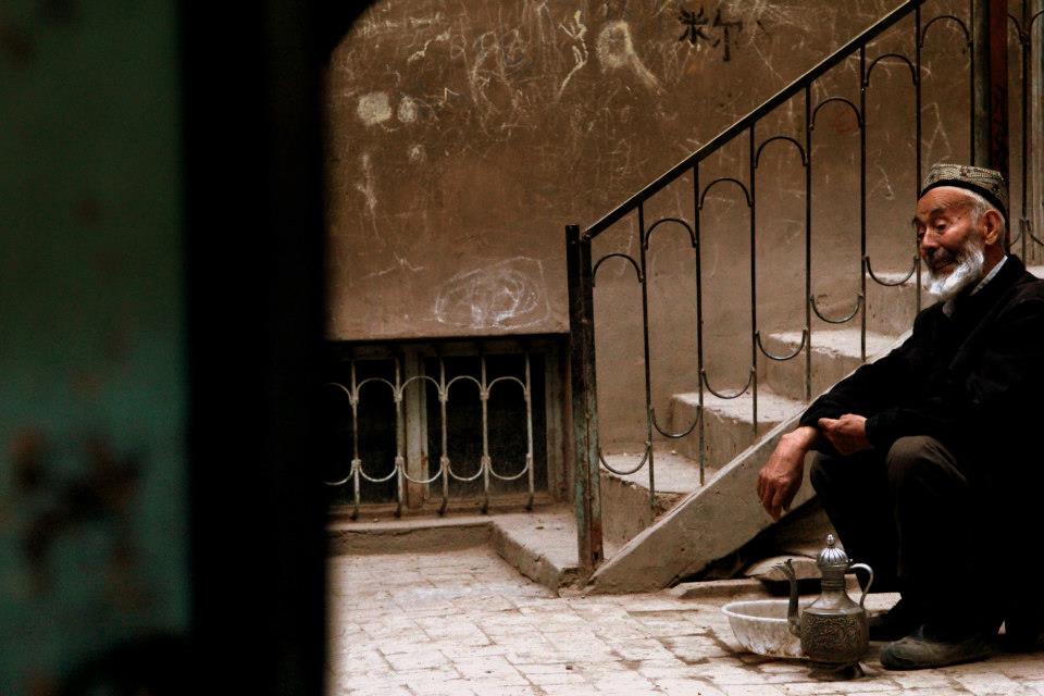 An elderly man with a white beard sits on a step beside a metal railing, wearing a traditional cap. He is outdoors in Xinjiang, amidst stunning landscapes and rustic charm, with a textured wall in the background, accompanied by a teapot and bowl on the ground.