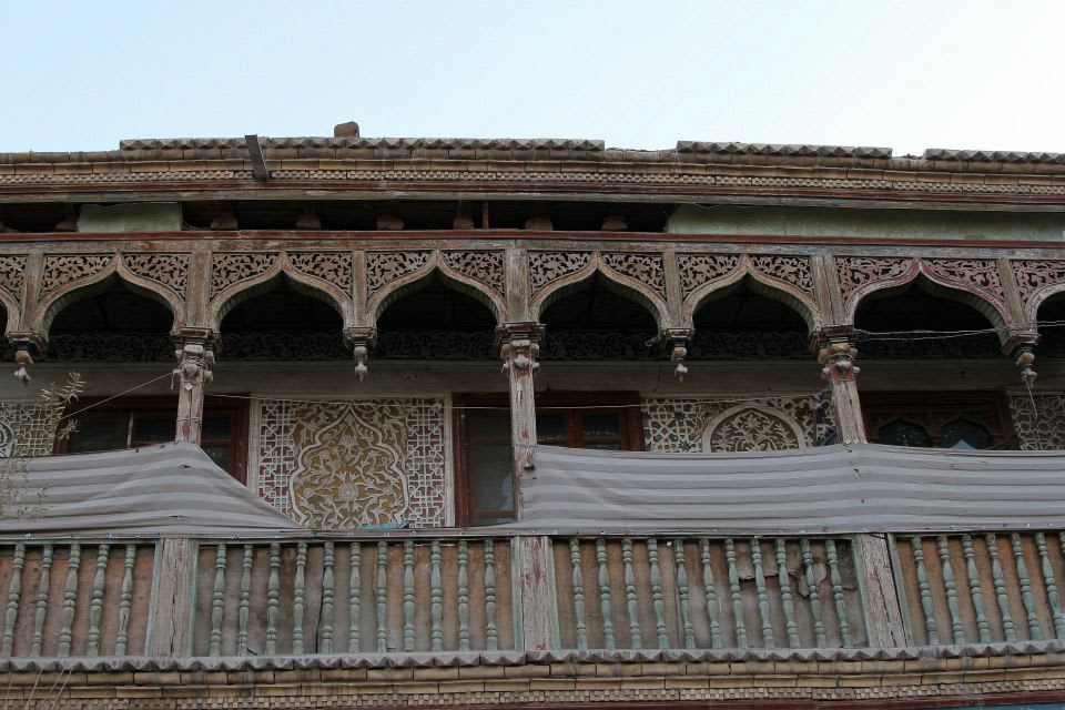 Rustic two-story building with intricate carved wooden arches and railings, reflecting Uyghur culture. The facade features ornate patterns and a wooden balcony partially covered with white fabric. This architecture captures the essence of Xinjiang's traditional styles amidst stunning landscapes.
