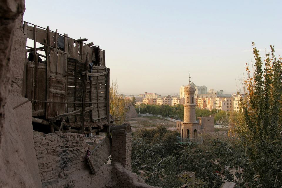 A scenic view of an old building in the foreground with wooden balconies captures the essence of Uyghur culture, overlooking a landscape that blends a traditional minaret and modern city buildings surrounded by trees. The stunning landscapes stand out under the clear daytime sky.