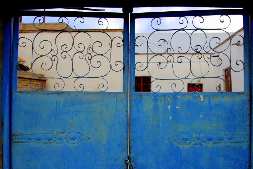 Blue wrought iron gate with intricate swirl designs, set against a backdrop of white buildings in the stunning landscapes of Xinjiang, under a clear sky.