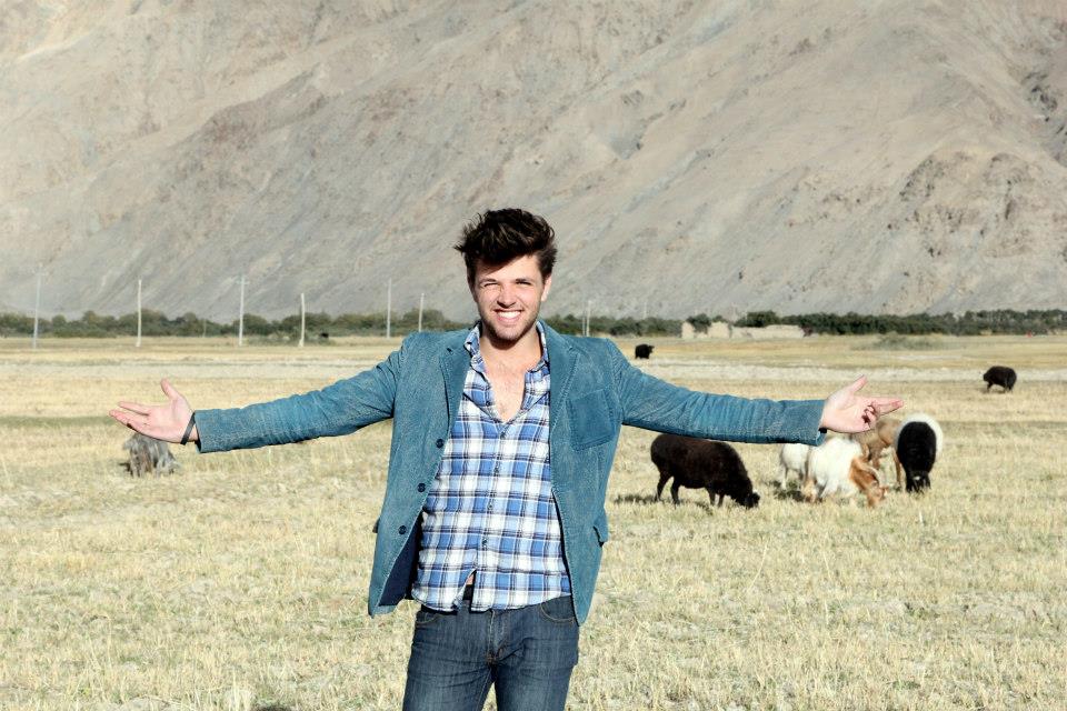 A man stands smiling with arms outstretched in a dry field, embracing the stunning landscapes of Xinjiang. He wears a blue jacket over a plaid shirt. In the background, several yaks graze, and a barren mountain range rises under a clear sky.