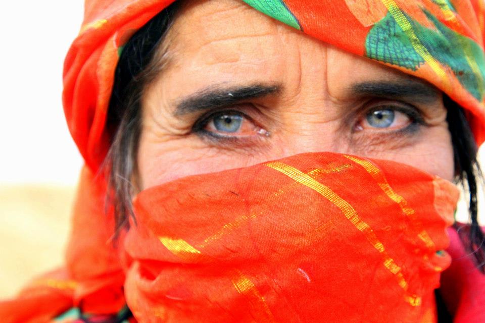 A person with striking blue eyes is wearing a vibrant orange headscarf and face covering, reflecting the rich Uyghur culture of Xinjiang. The scarf has green and gold accents, highlighting their expressive gaze.