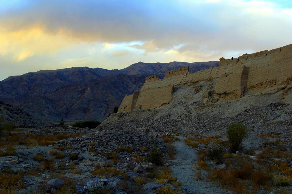 A rugged pathway winds through a rocky landscape with large, eroded cliffs on the right. In Xinjiang's stunning landscapes, distant mountains are silhouetted against a cloudy sky, bathed in warm hues from a setting sun. Sparse vegetation dots the foreground.