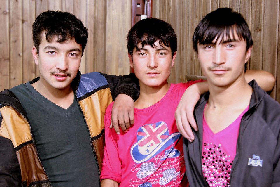 Three young men stand together indoors, each with dark hair. The man on the left wears a jacket, the middle one a pink t-shirt with a graphic, and the one on the right a dark jacket over a bright pink shirt. Their close pose exudes camaraderie reminiscent of vibrant Xinjiang culture.