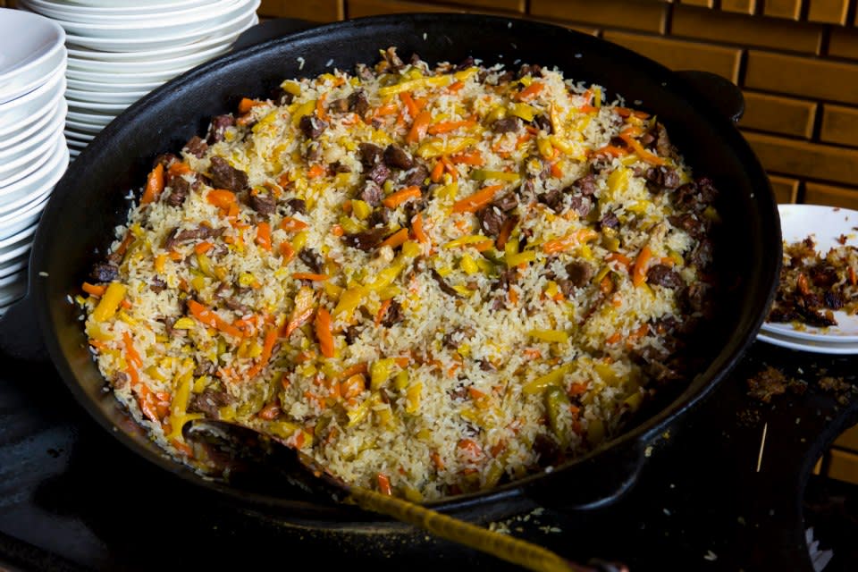 A large pan filled with a colorful rice dish, evoking the rich flavors of Uyghur culture, contains pieces of meat, carrots, and peppers. A serving spoon rests on the side. Stacks of white plates are visible in the background on the left, reminiscent of street feasts found in stunning Xinjiang landscapes.
