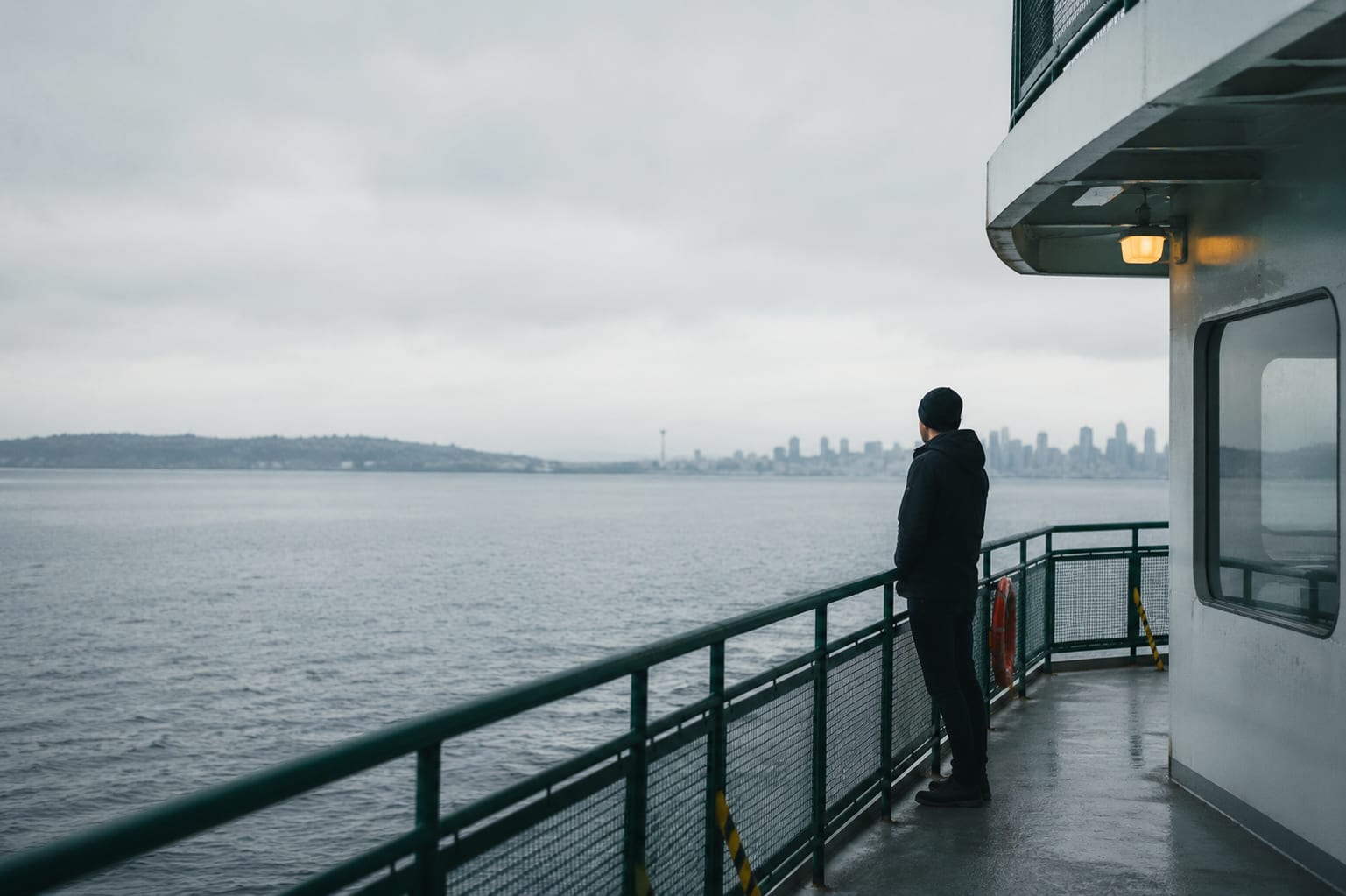 A solitary figure standing on a ferry deck, looking toward the Seattle skyline under a gray overcast sky.