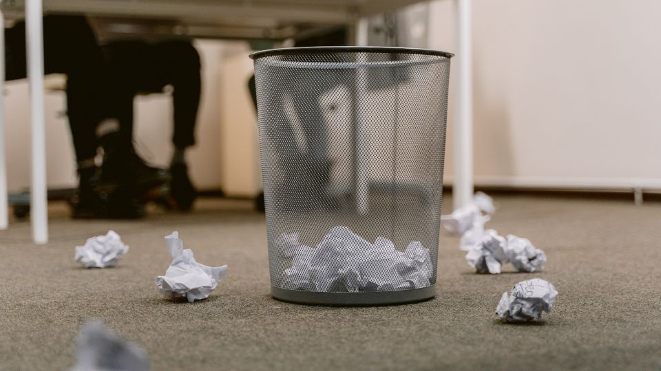 a photograph of a paper waste bin in an office. In the foreground you can see a paper waste basket made of a metal mesh material and lots of scrumpled up balls of paper in and around the basket. In the background you can see a desk and some feet with office wear on.