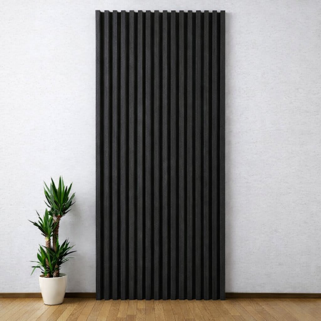 A tall, vertical WPC wall panel with dark gray textured slats stands out against a light-colored wall in a minimalist, modern interior, paired with a small potted plant on a warm wooden floor, creating a serene and clean design.
