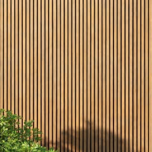 WPC slatted wall panels in warm tones, featuring uniform vertical slats and a natural wood grain texture, framed by green foliage and soft shadows. A minimalist architectural facade ideal for backgrounds, exteriors, design, and nature-inspired concepts.